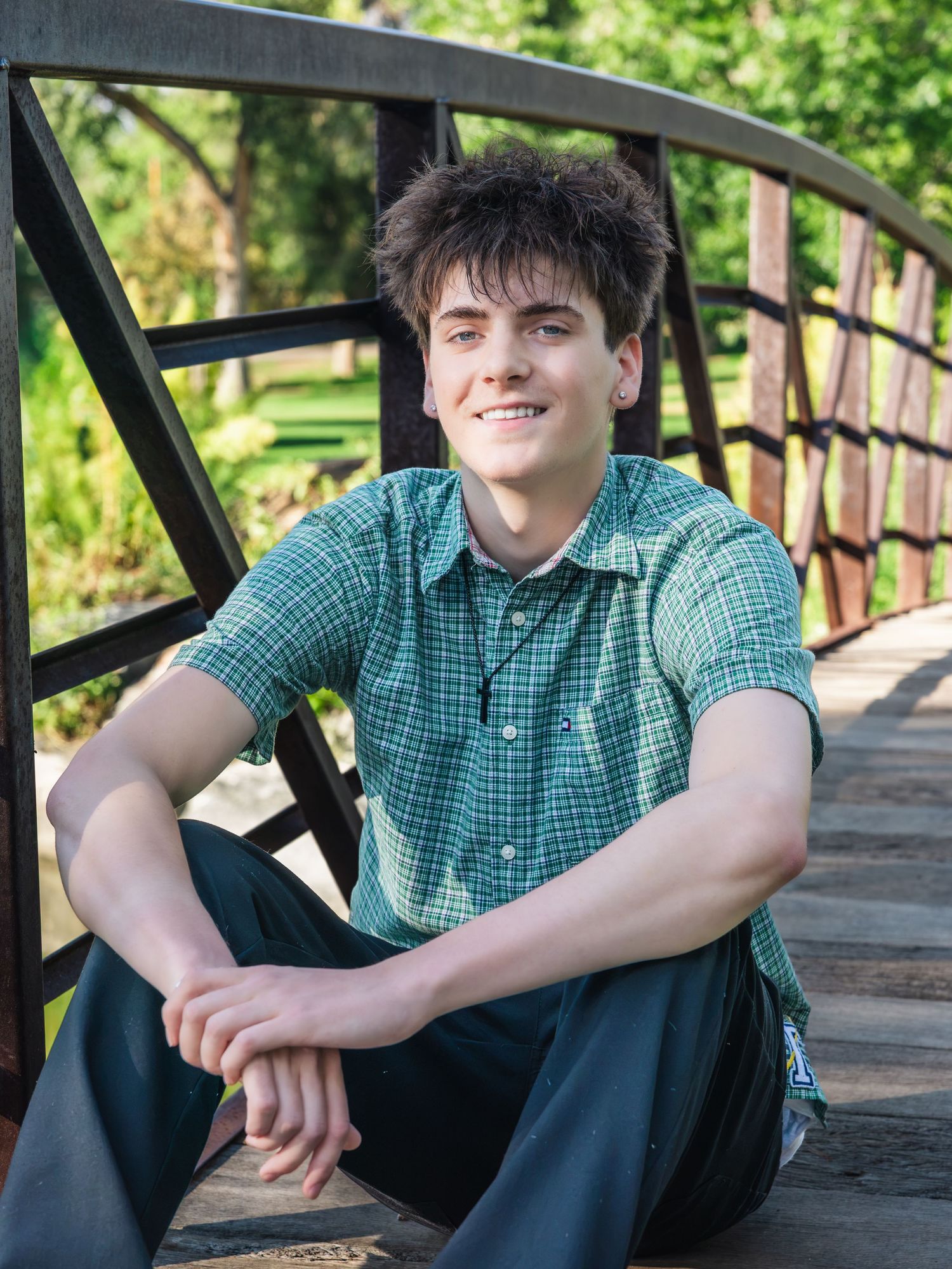 Smiling teenage boy in green plaid shirt sitting on wooden bridge steps in a sunny park setting.