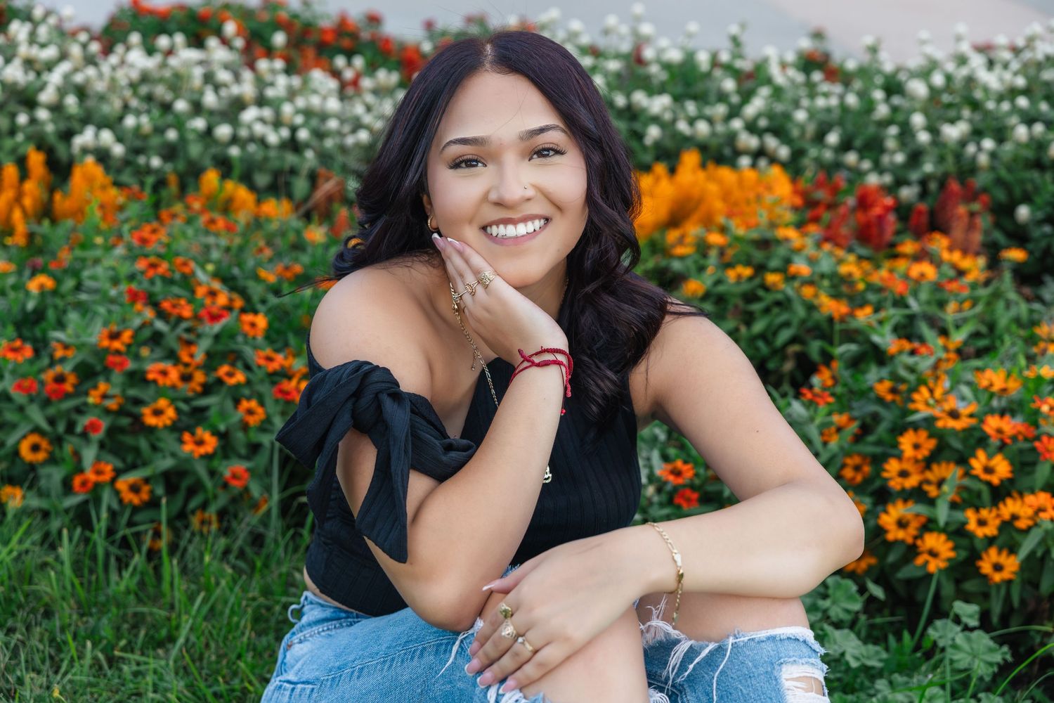 Young woman smiling in front of colorful flower garden, wearing black top and denim shorts.
