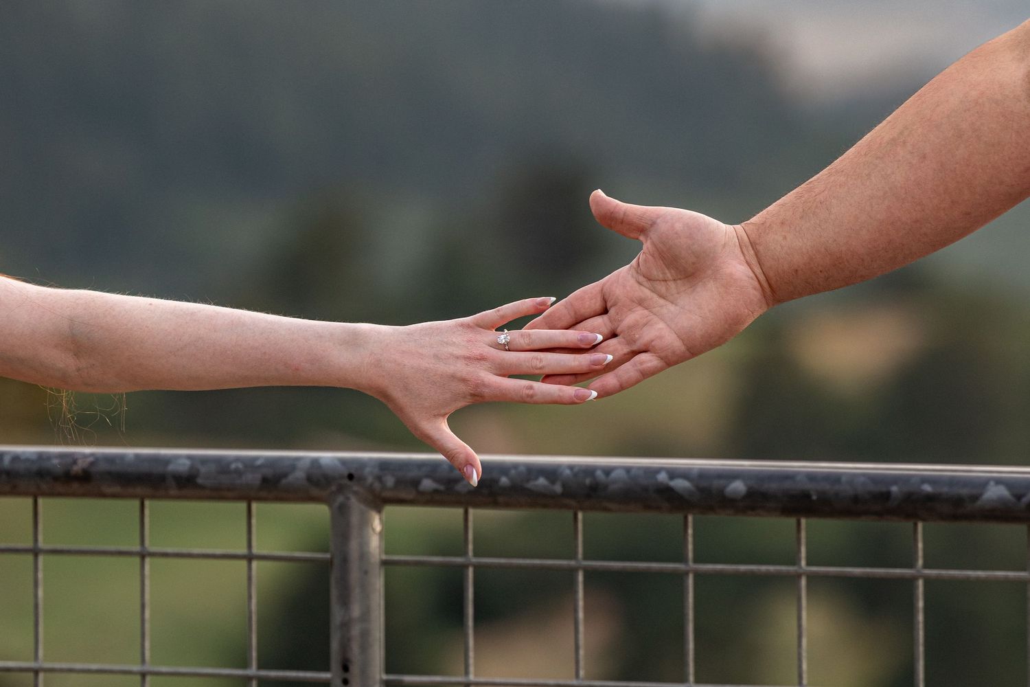 Two hands reaching toward each other over a metal railing with a blurred green mountain landscape in the background.