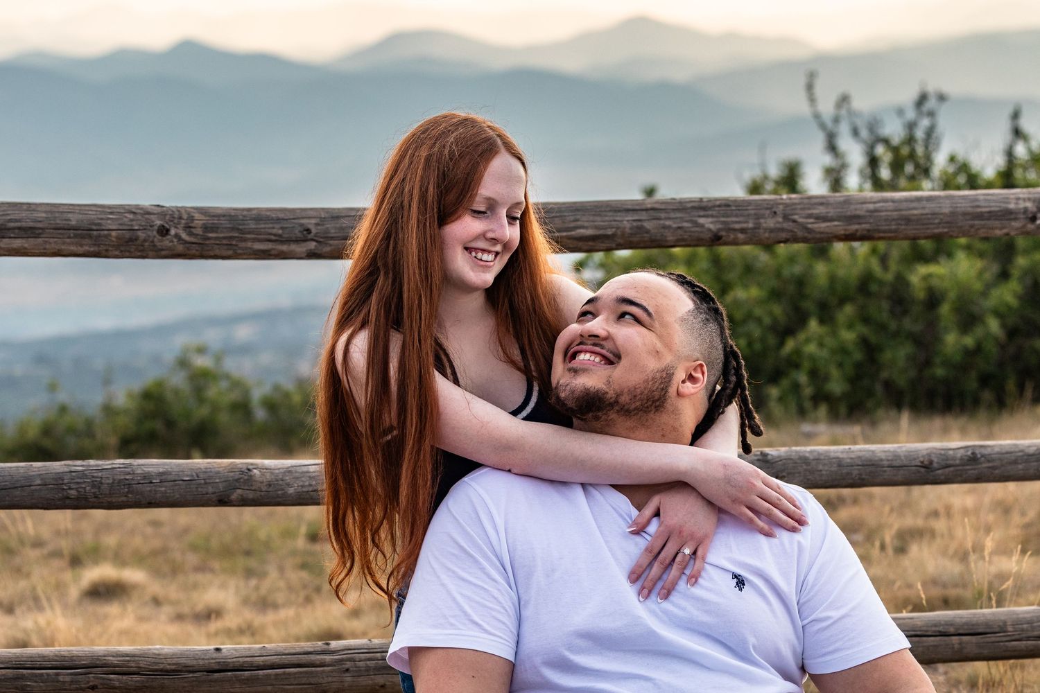 A smiling couple poses outdoors on a bench with mountain views, woman with long red hair embracing a man in a white shirt.