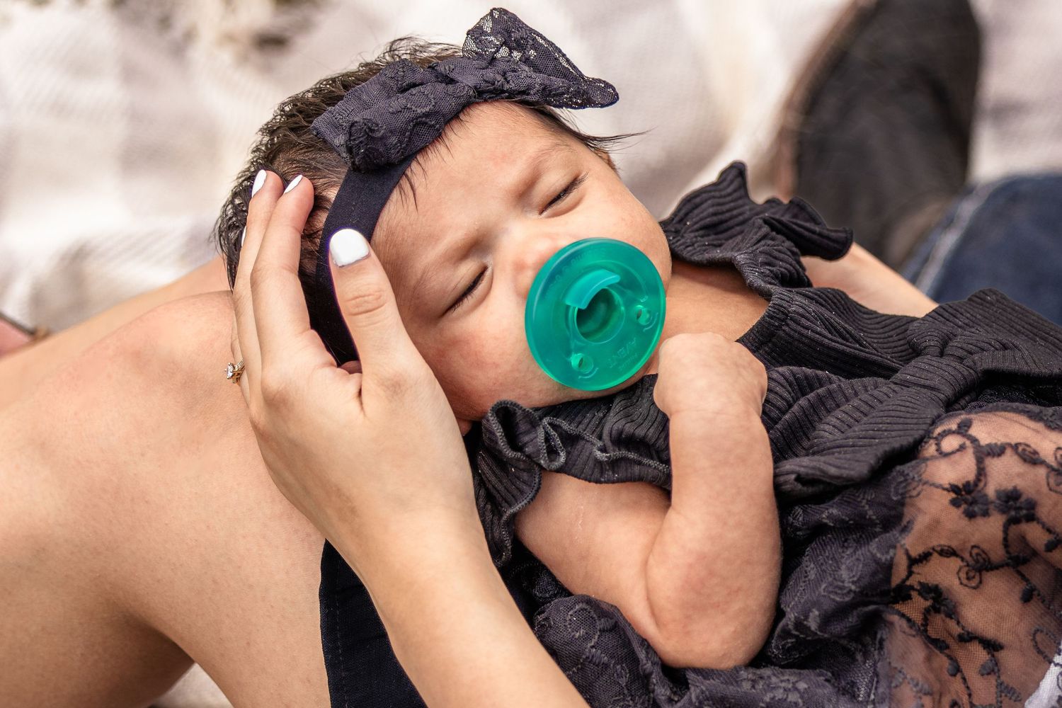 A mother cradles her sleeping newborn baby with a teal pacifier and black lace outfit and bow headband.