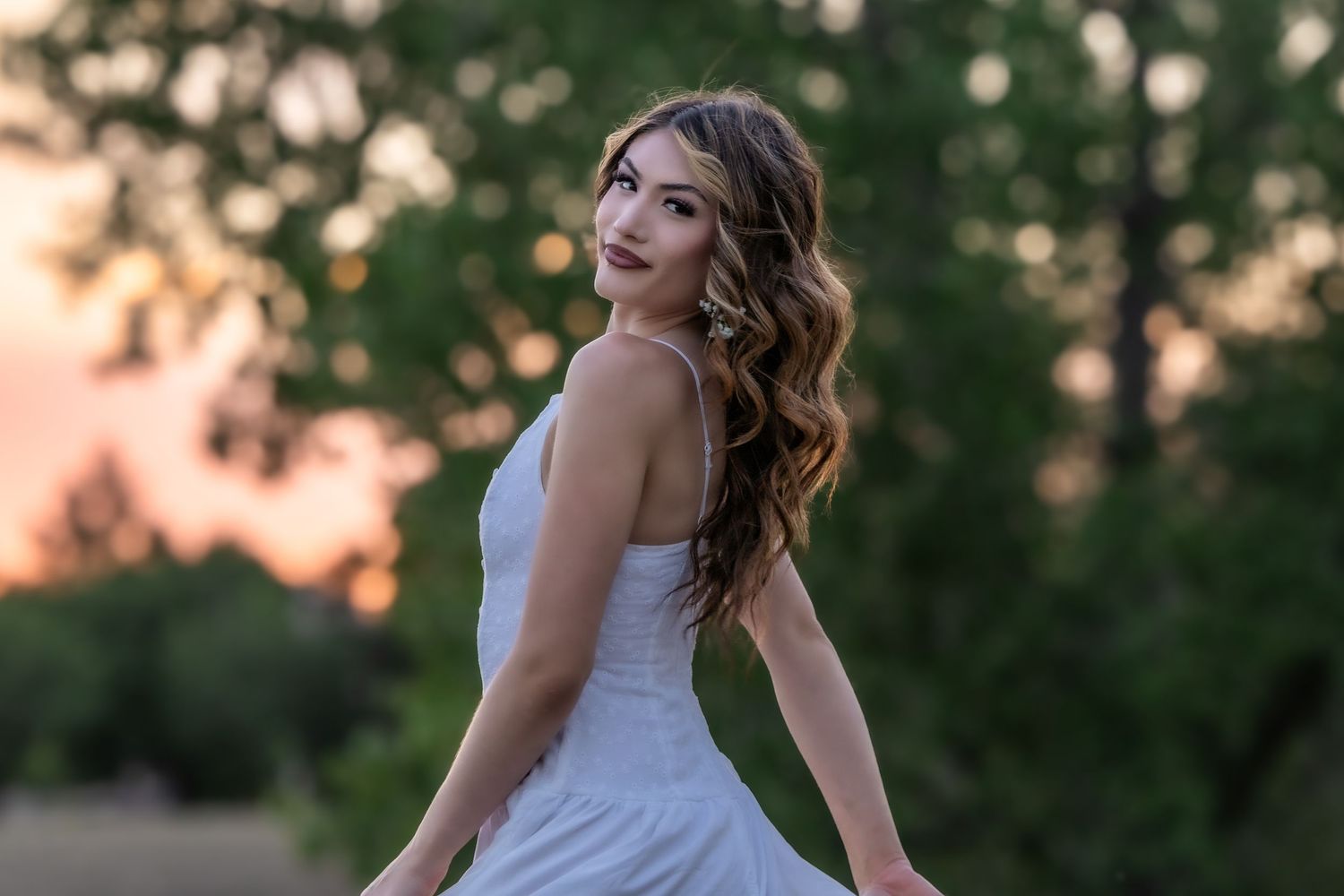 Young woman in white dress with curly hair smiles over shoulder at golden sunset in Littleton, Colorado.