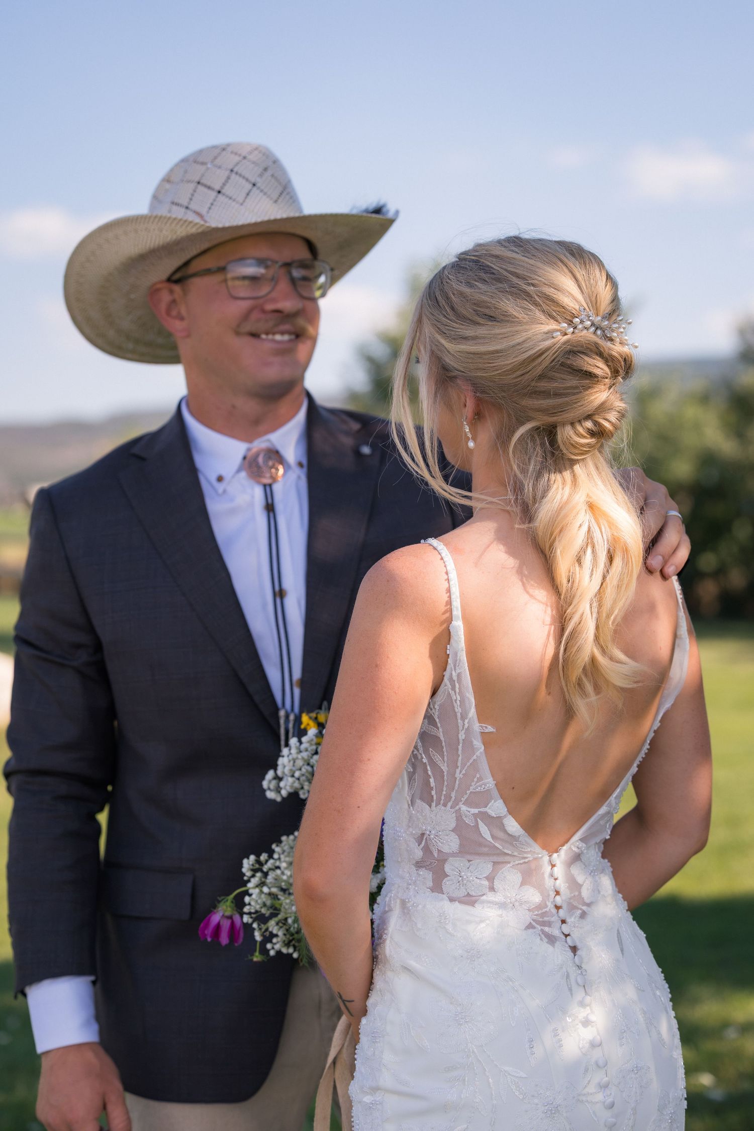 A smiling groom in a cowboy hat and bolo tie stands beside a bride in a backless lace wedding gown outdoors.