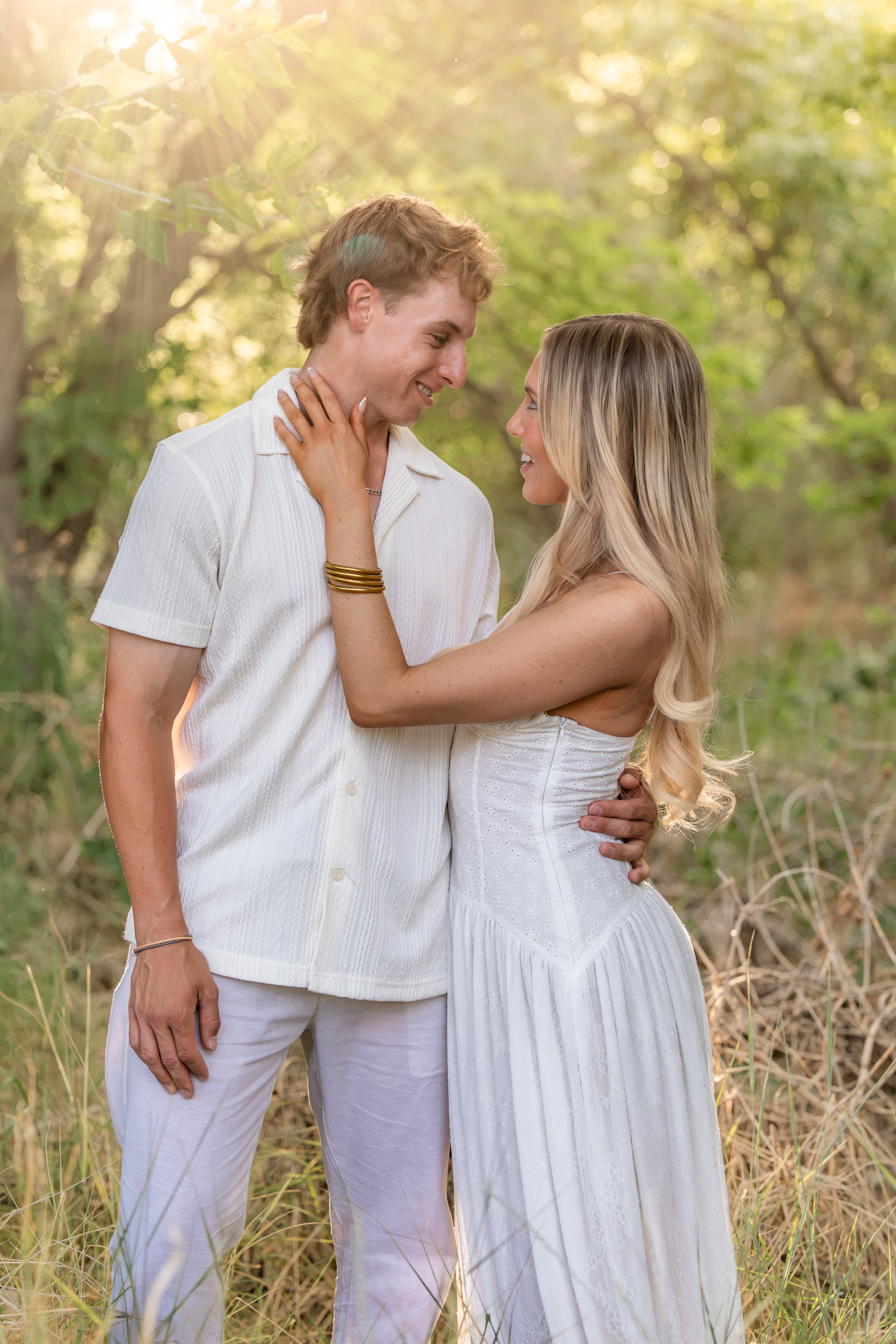 A couple in matching white outfits embrace and smile at each other in a sunlit outdoor setting.