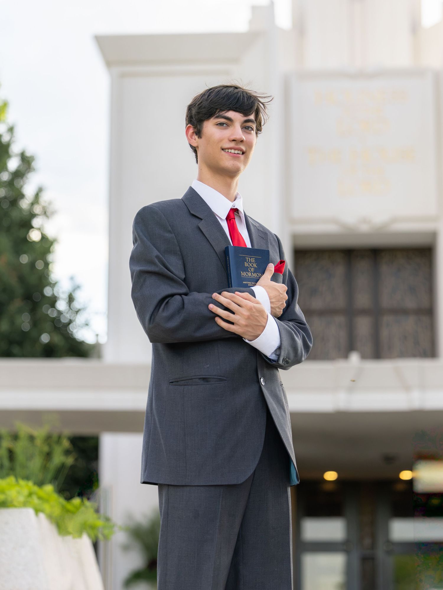 Young man in gray suit and red tie holding a book, smiling outdoors near a white building.