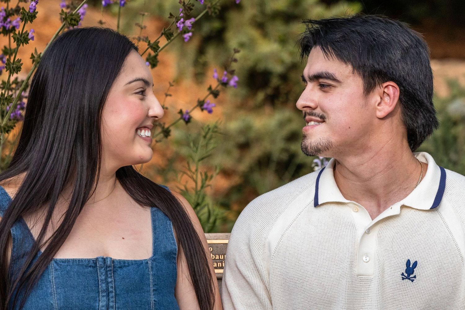 A young couple smiling at each other outdoors, surrounded by purple flowers and greenery.
