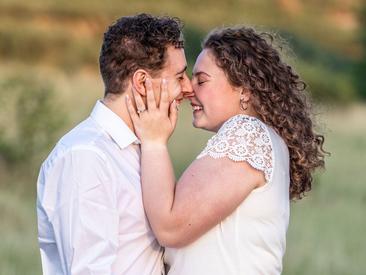 Couple sharing a romantic moment outdoors, woman smiling and holding man's face, wearing white outfits in a green field.