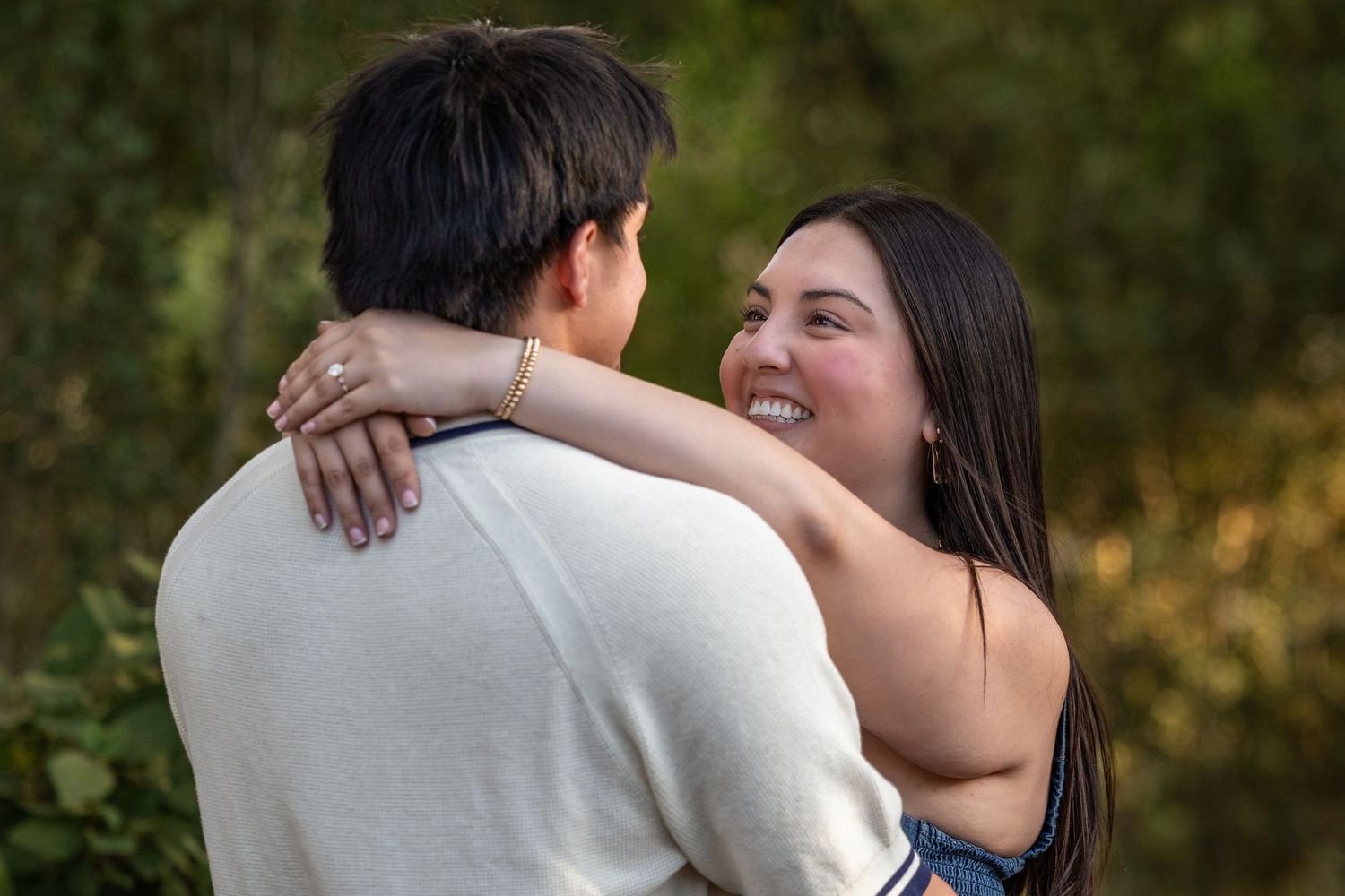 A smiling young woman embraces a man outdoors, with lush green foliage in the background.