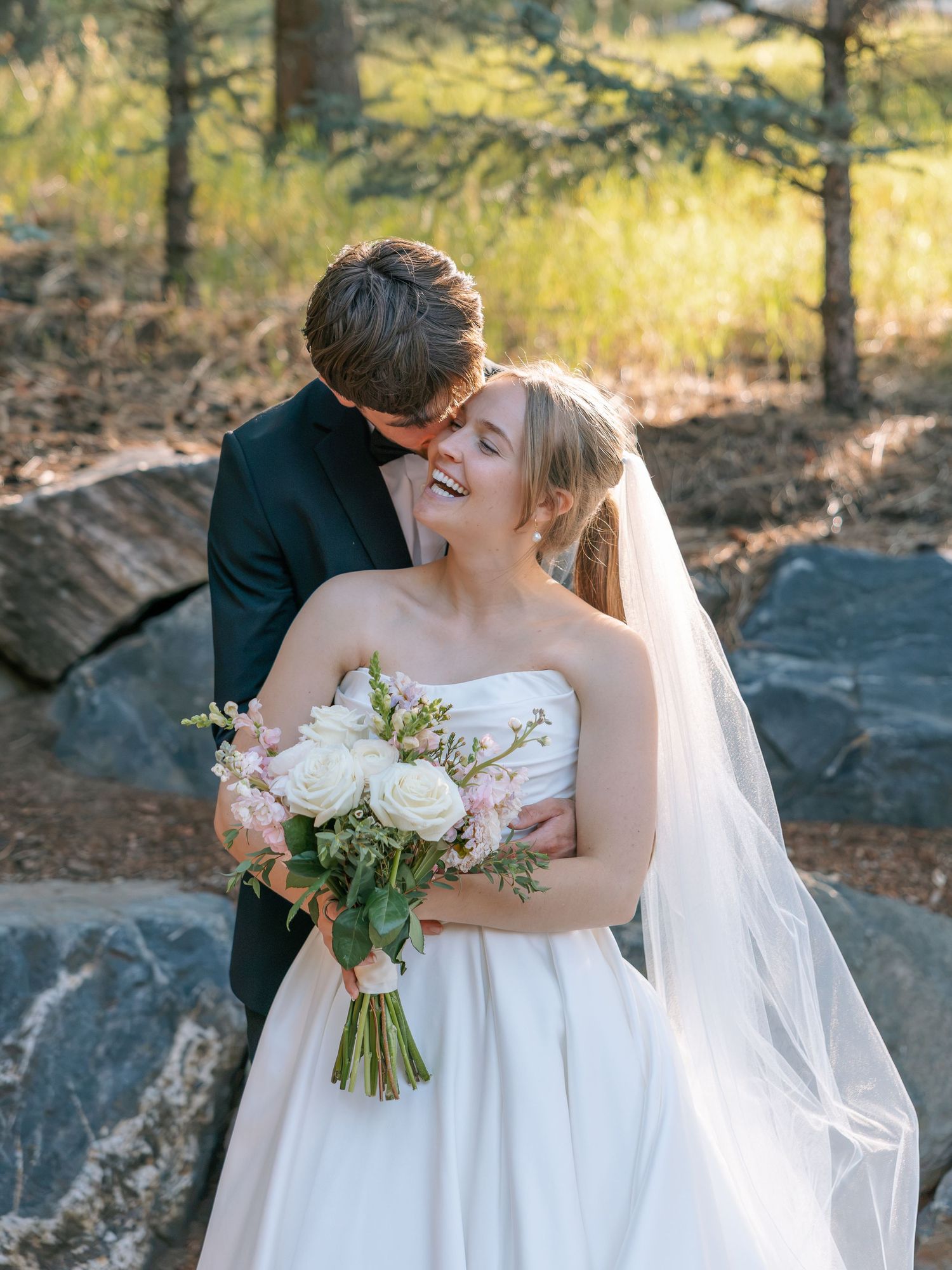 Smiling bride in strapless gown holds white bouquet as groom embraces her near rustic stone wall.