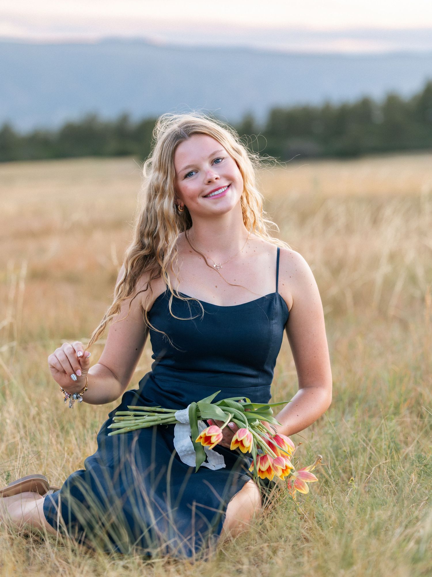 Young blonde woman in black dress sitting in golden field holding colorful tulips with mountains in background.