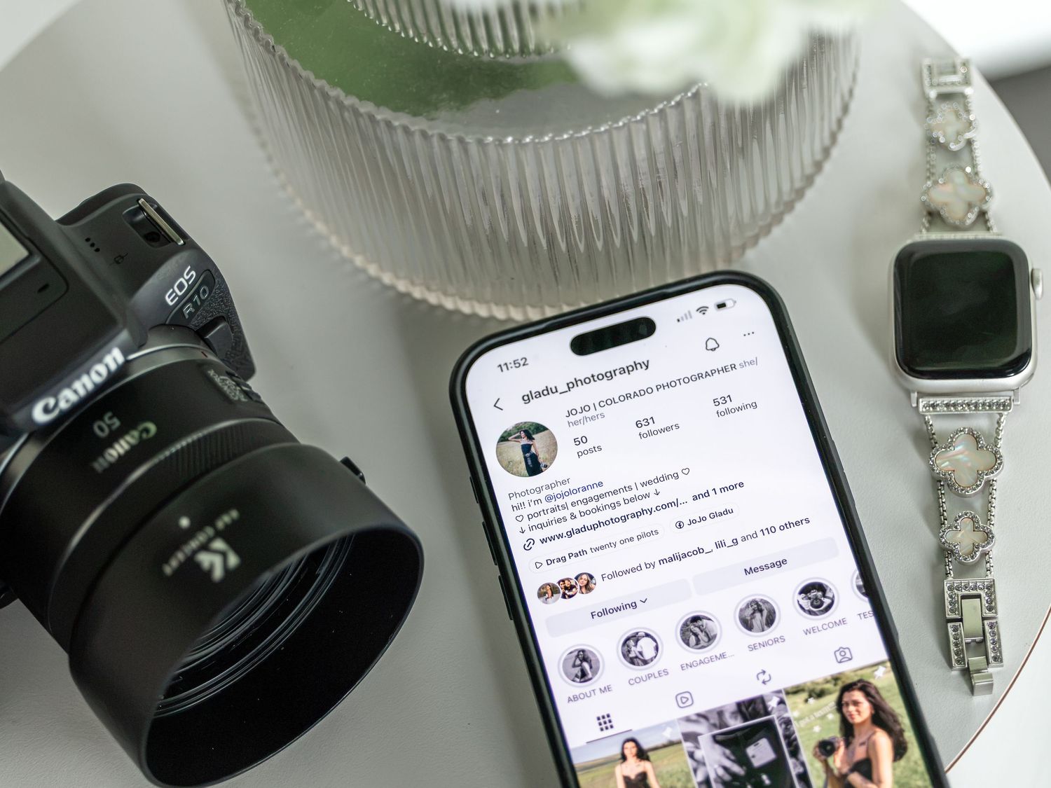 Canon DSLR camera beside iPhone showing Instagram profile, silver Apple Watch on white table.
