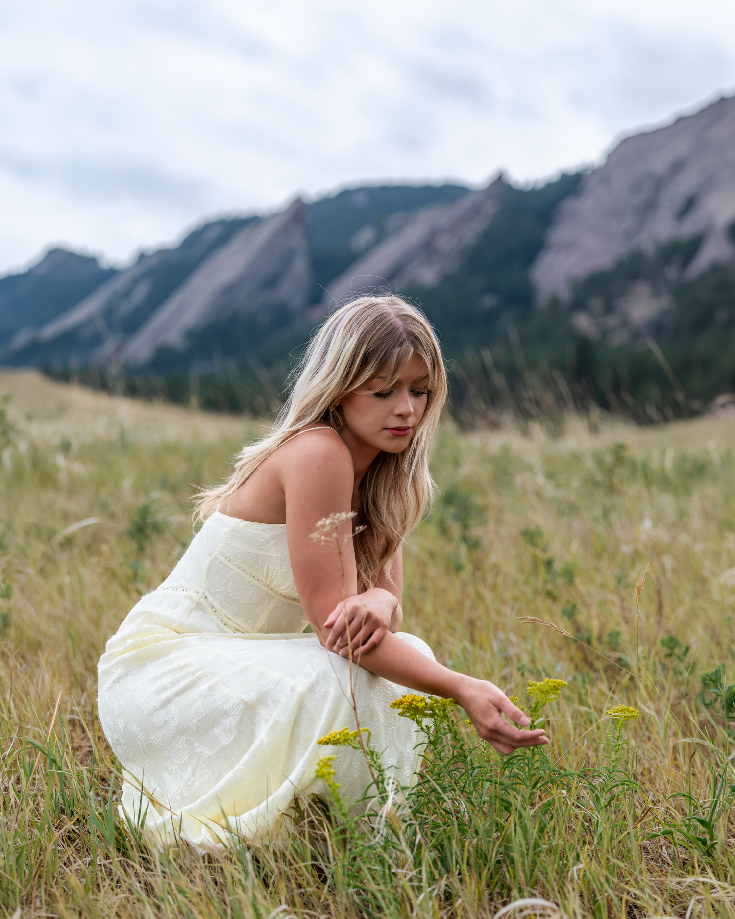 A blonde woman in a white dress crouches in a mountain meadow, gently touching wildflowers with the Flatirons in the background.