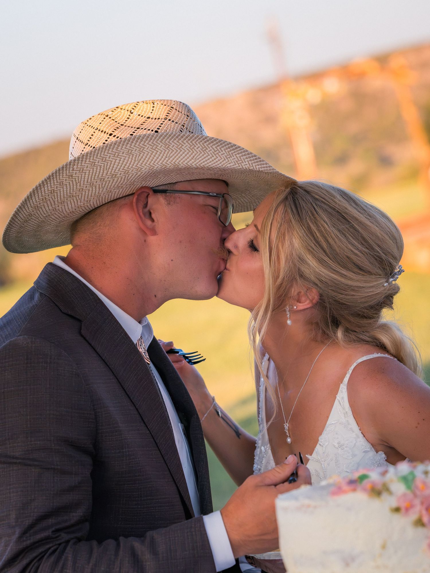 Newlyweds share a romantic kiss at cake cutting, groom wearing cowboy hat at outdoor wedding reception.