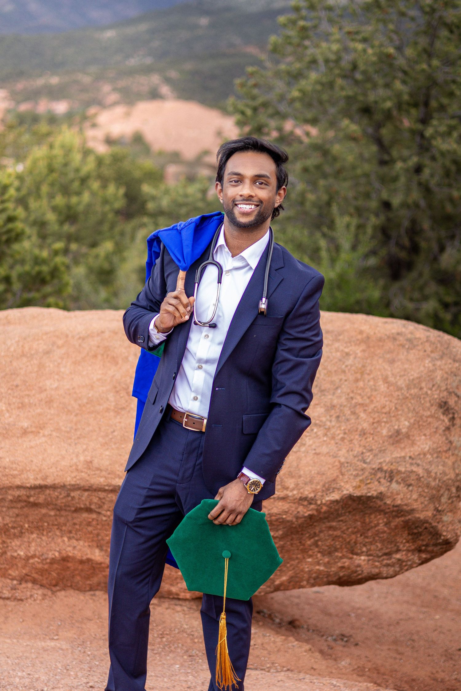 Smiling medical graduate in navy suit with stethoscope, blue gown, and green cap poses outdoors near red rocks.
