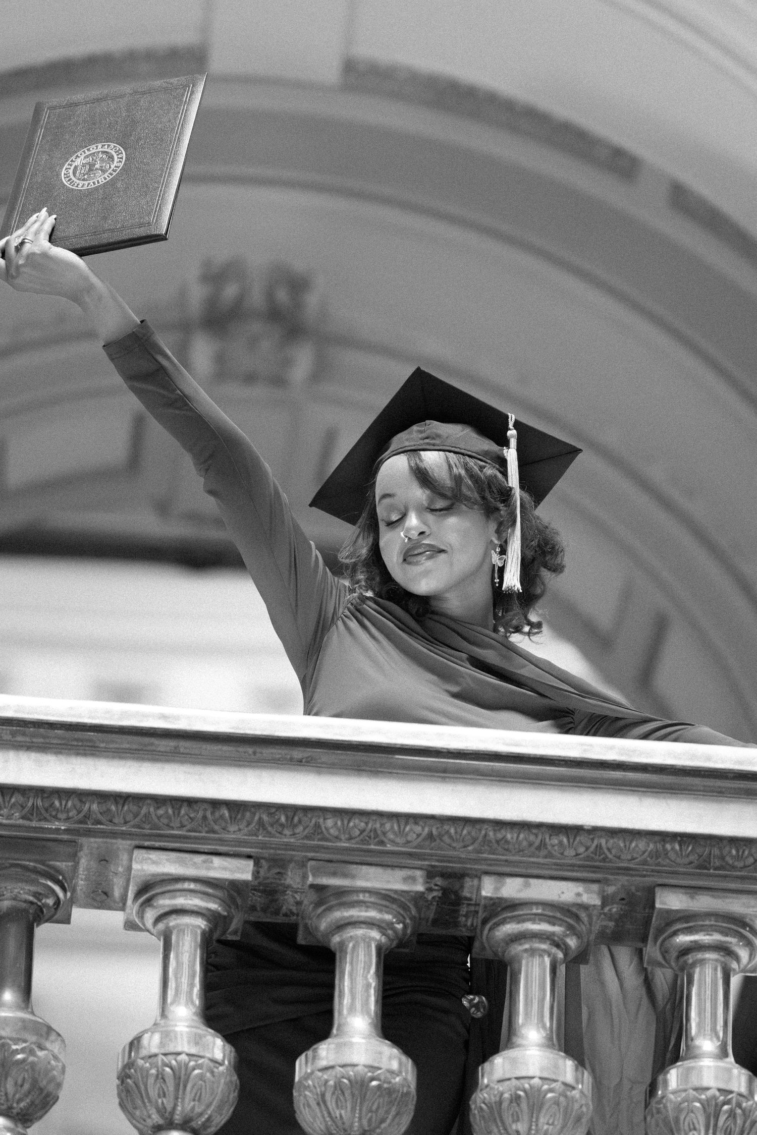 Black and white photo of graduate raising diploma on ornate balcony wearing mortarboard cap in Littleton, Colorado.