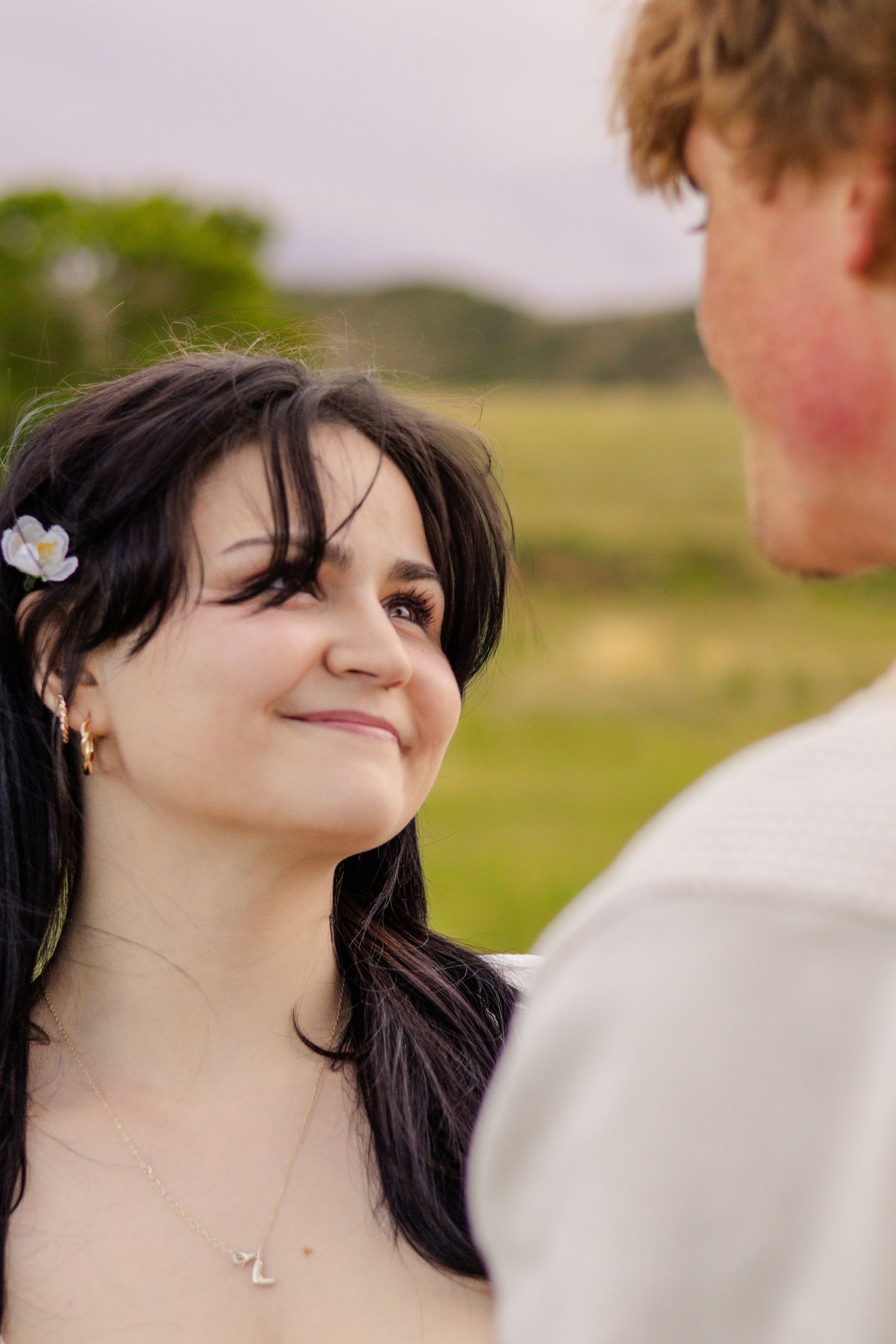 A dark-haired woman with a flower in her hair smiles warmly at her partner in an outdoor field setting in Littleton, Colorado.