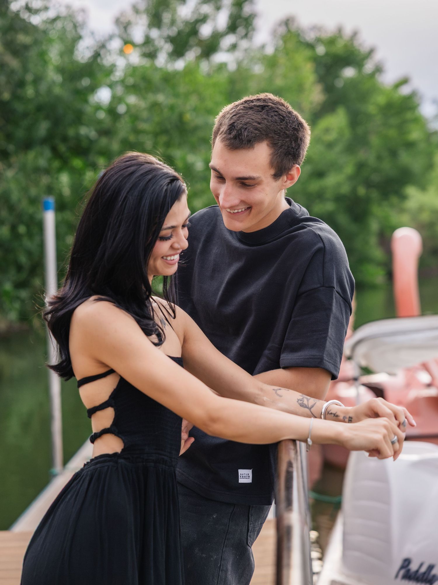 A smiling couple in black outfits share a romantic moment on a boat dock surrounded by lush green trees.