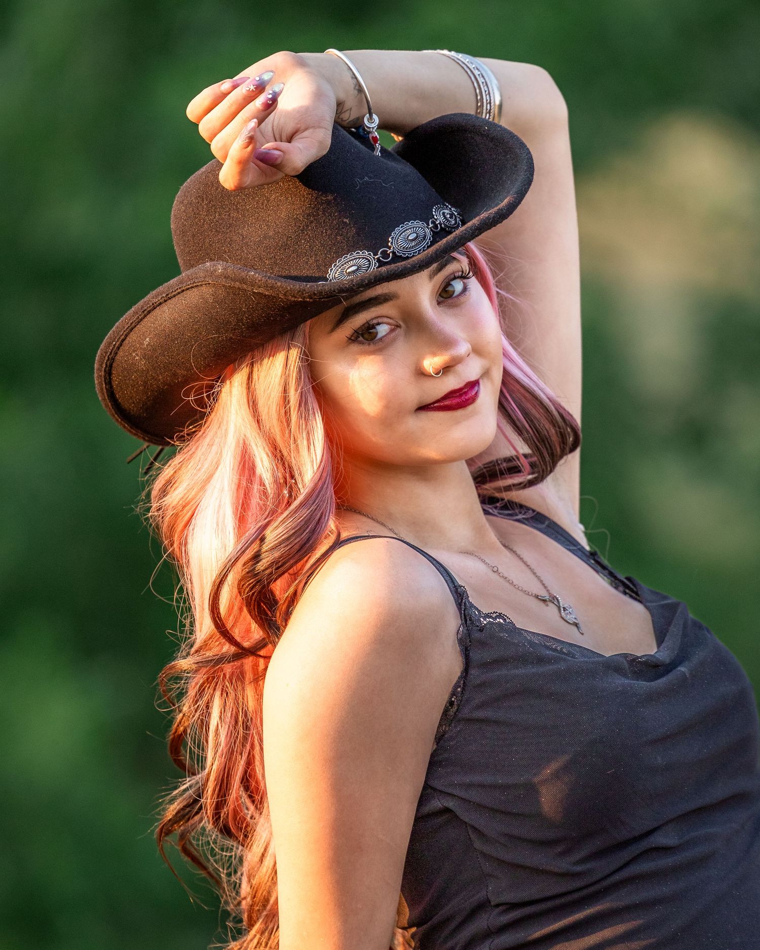 Young woman with pink hair holding a brown cowboy hat, wearing a gray top outdoors.