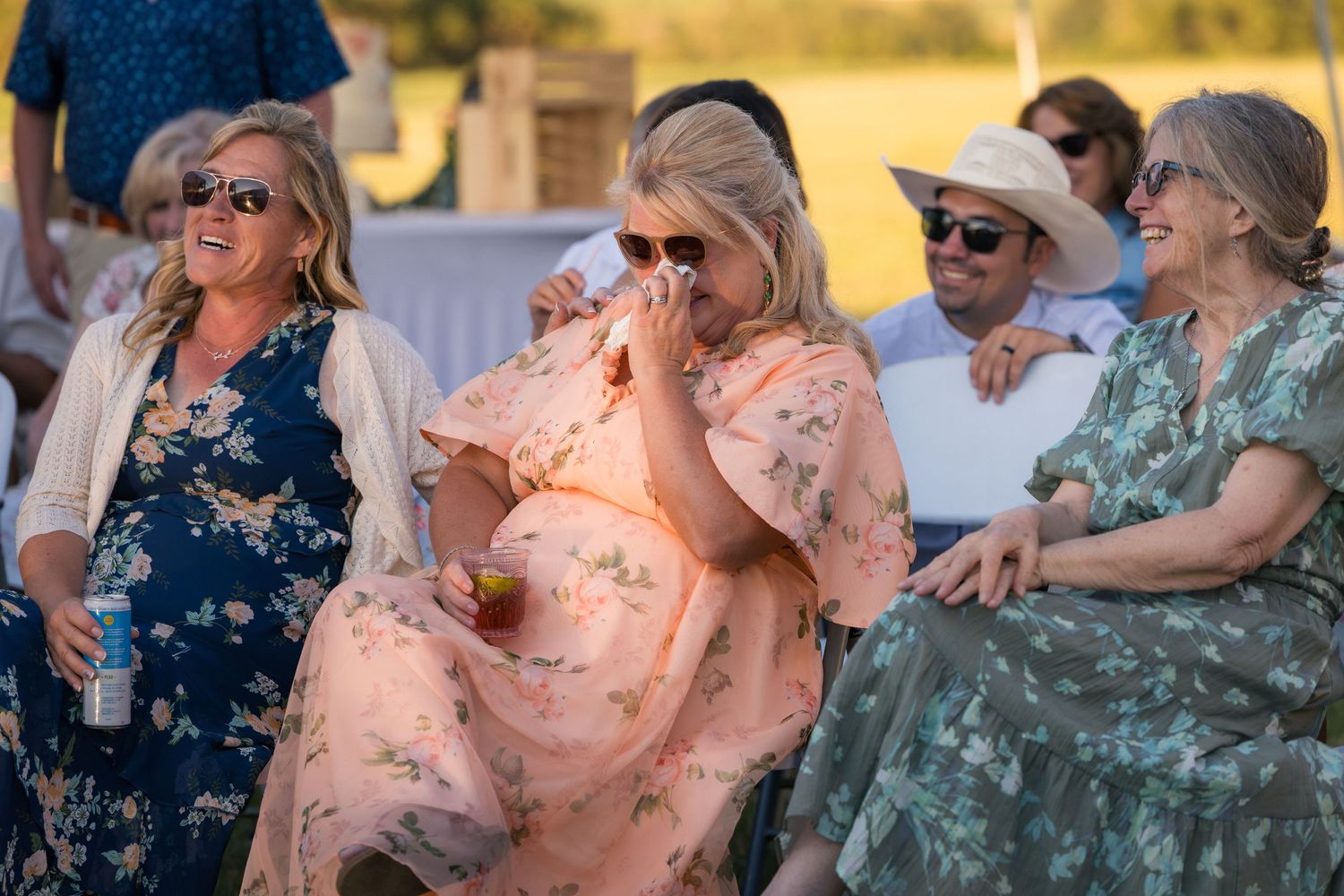 Women in floral dresses laughing and enjoying an outdoor wedding reception on a sunny day.