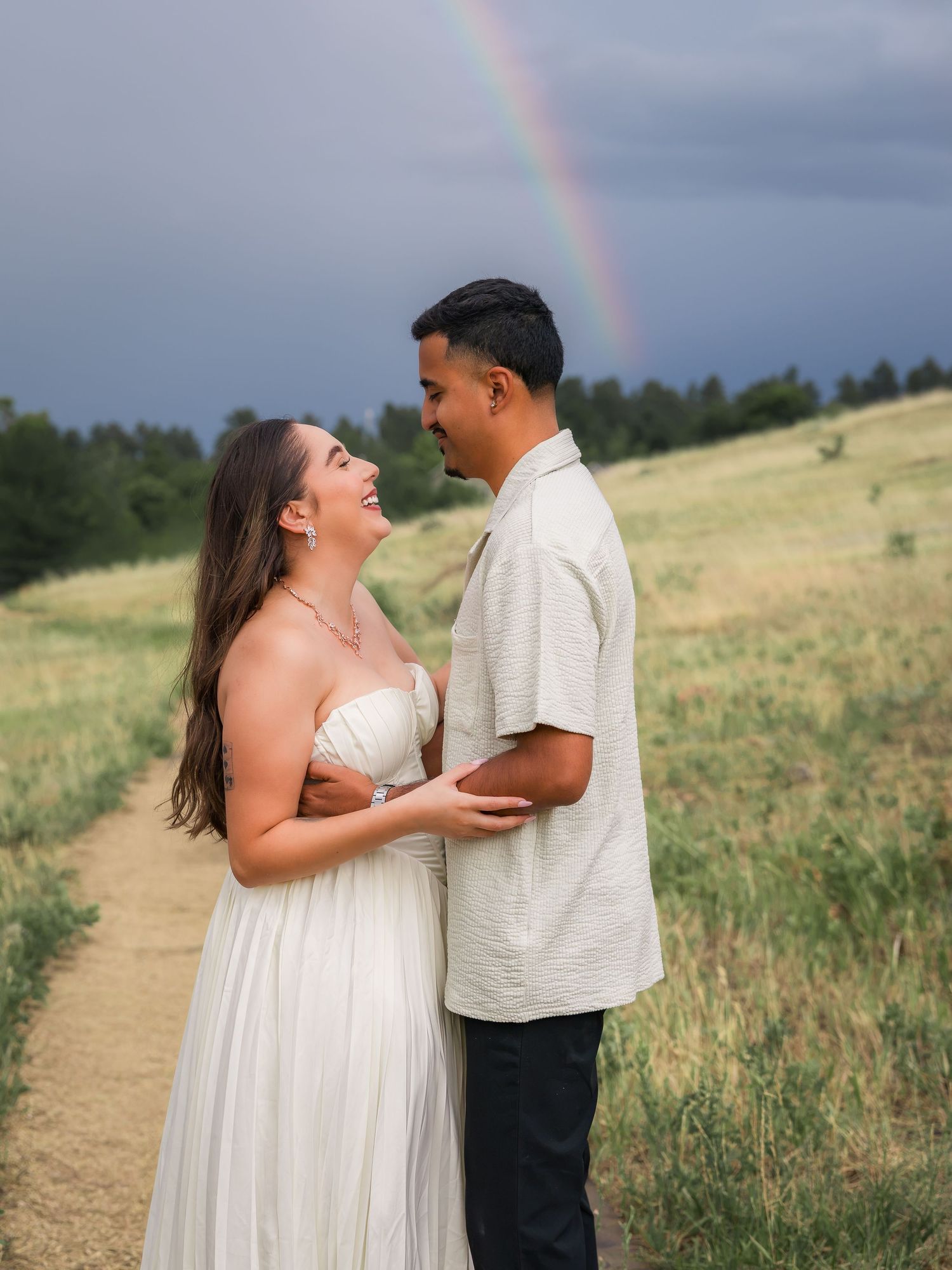 A couple shares a joyful moment on a scenic trail with a rainbow arching over green hills under a dramatic sky.