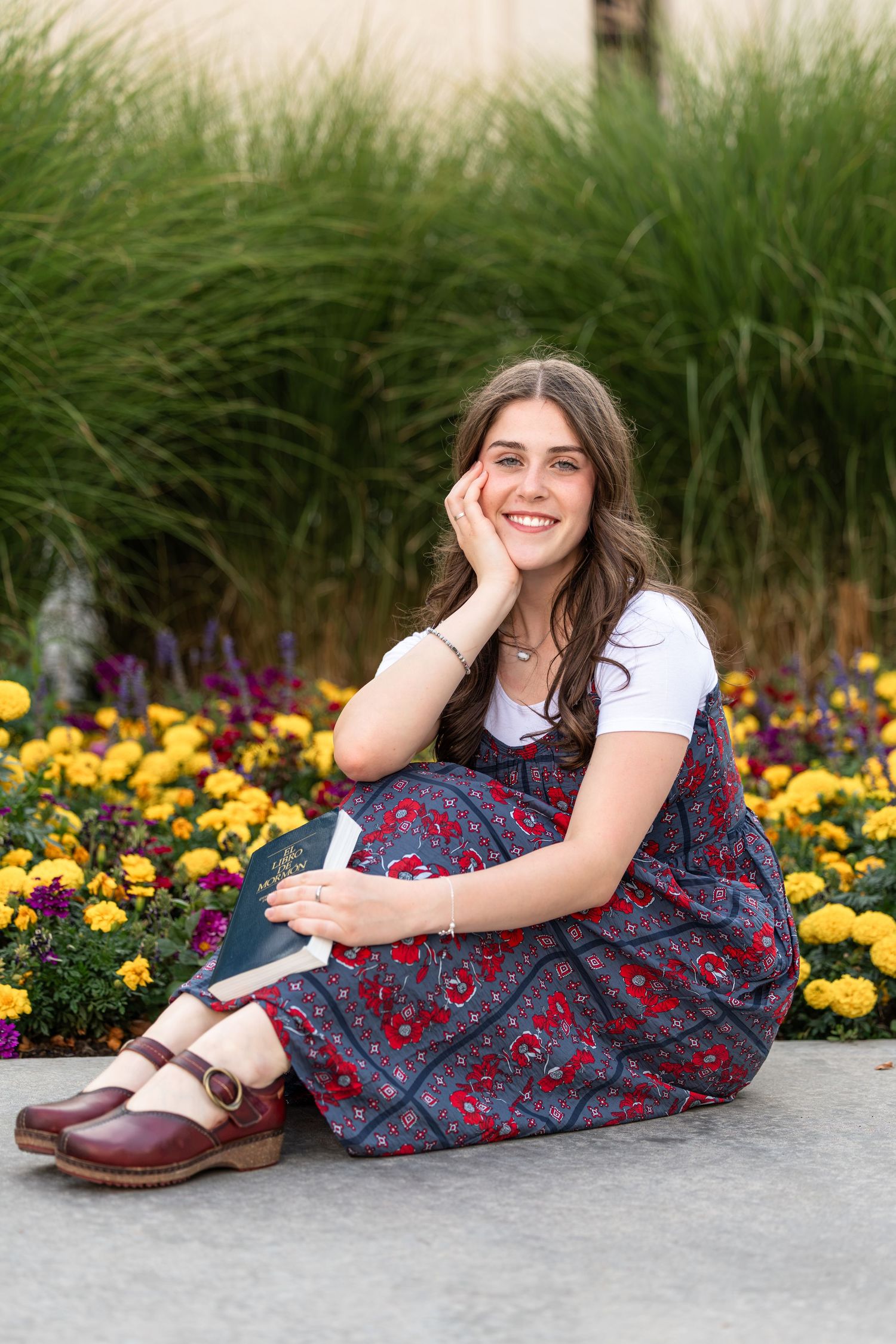 Young woman in floral dress sitting by colorful garden flowers, smiling with hand on cheek.