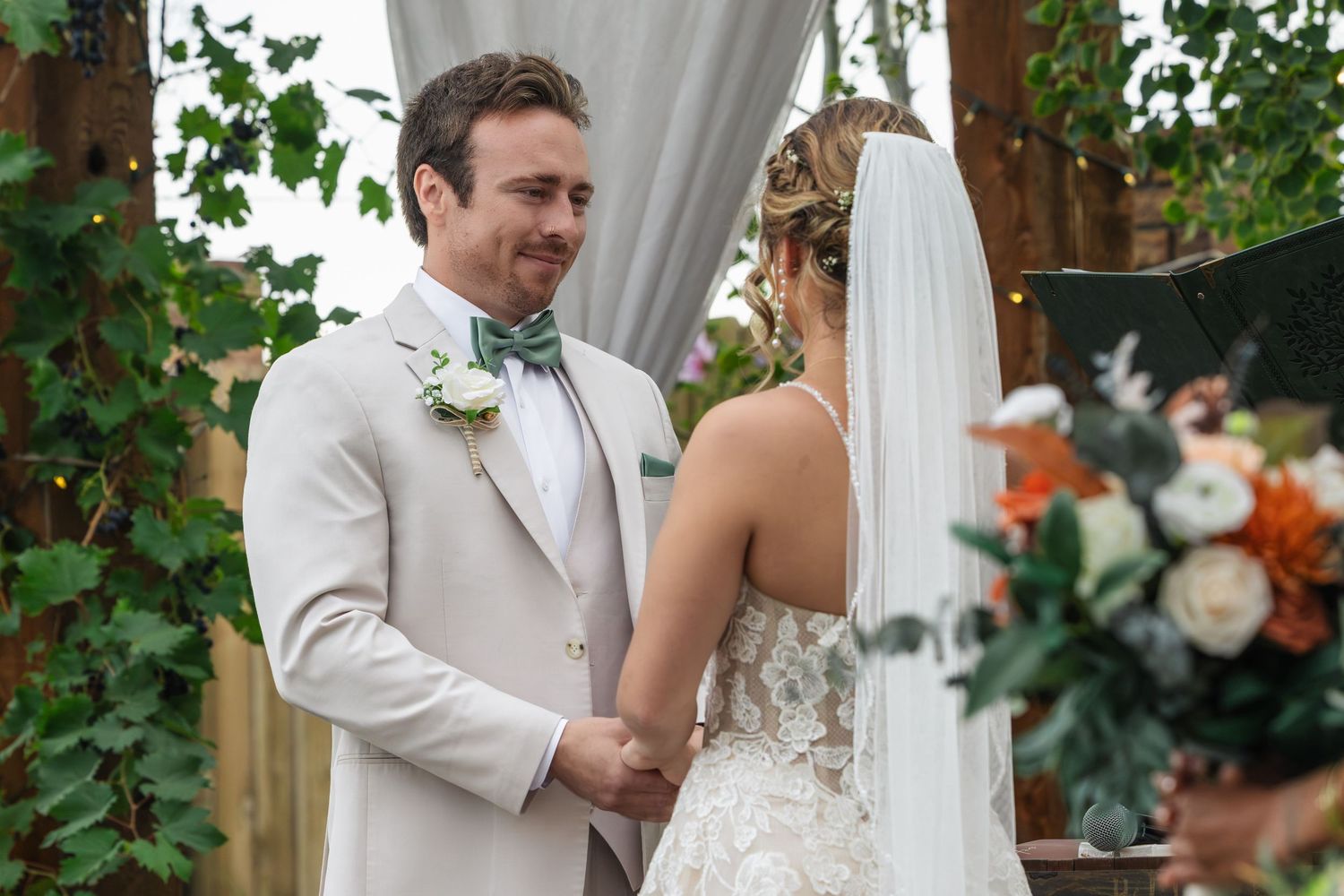 Bride and groom holding hands at outdoor wedding ceremony, groom in ivory suit with sage bow tie and boutonniere.