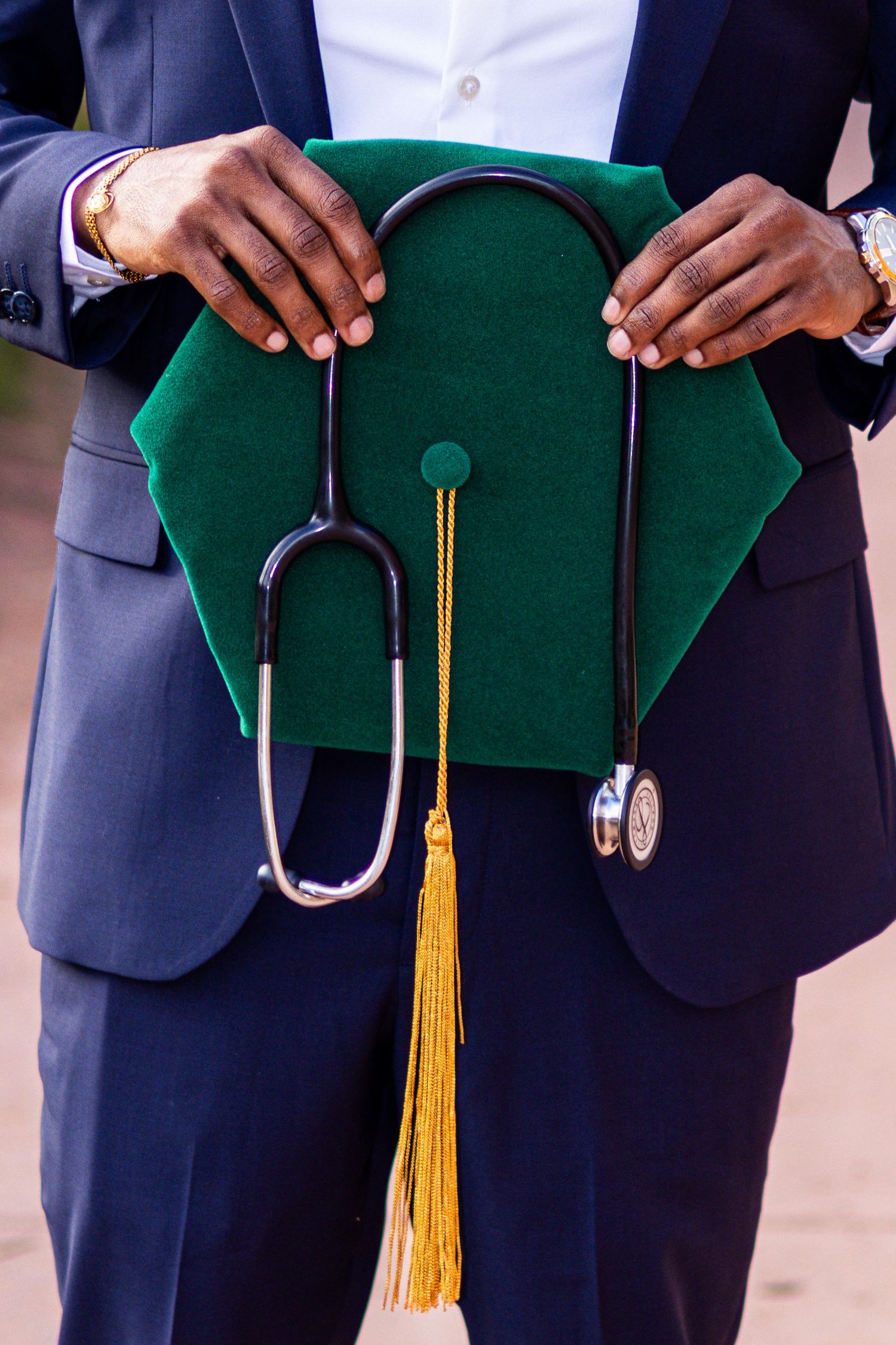 Graduate in navy suit holds green mortarboard with gold tassel and stethoscope draped over it.