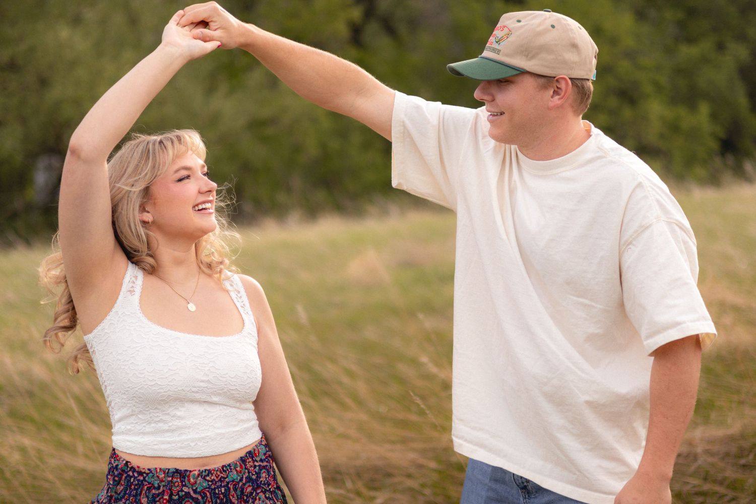 Smiling couple dancing outdoors in a field, man in white tee and cap spinning blonde woman in white top and floral skirt.