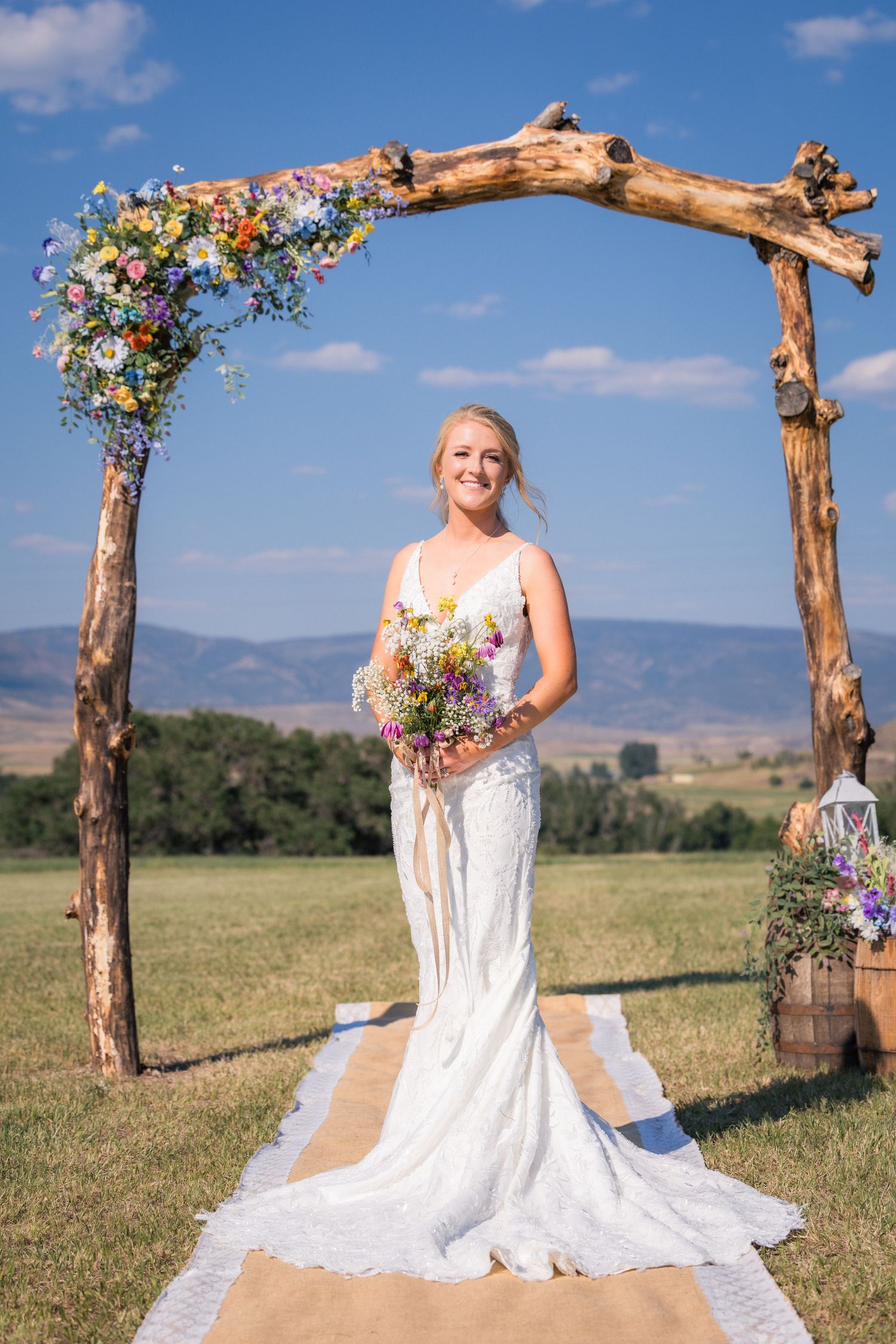 Smiling bride in white lace gown holding wildflower bouquet under rustic wooden arch with mountain backdrop.