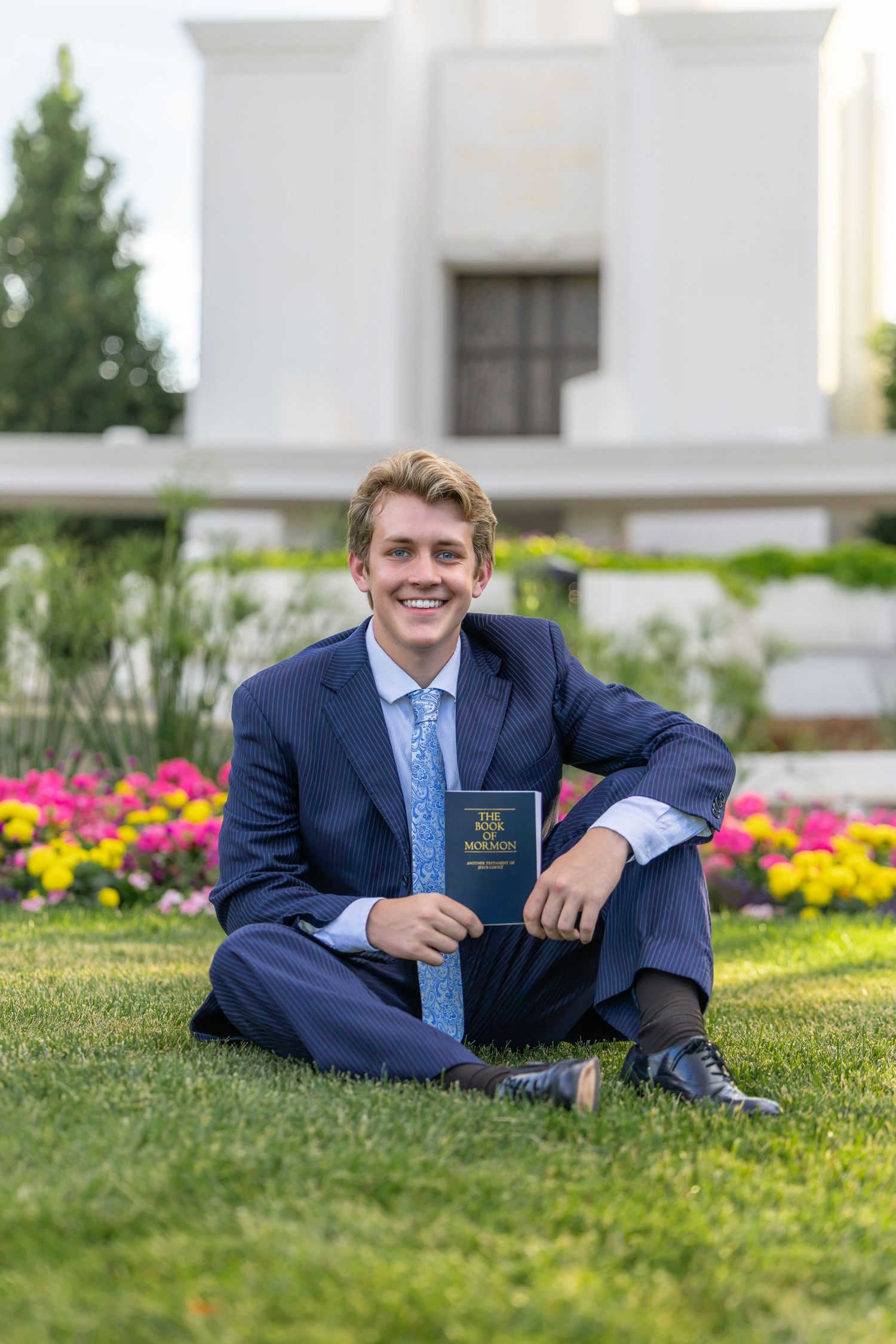 Young man in navy suit holding Book of Mormon, sitting on grass near flowering garden outside LDS temple.