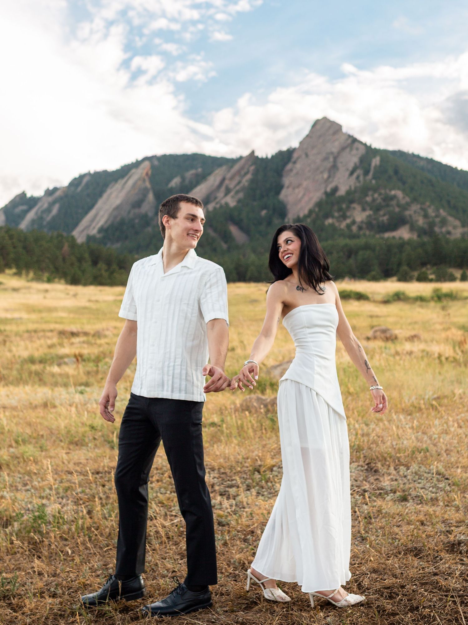 Couple in white outfits holding hands in a golden meadow with the iconic Flatirons mountains in Boulder, Colorado.