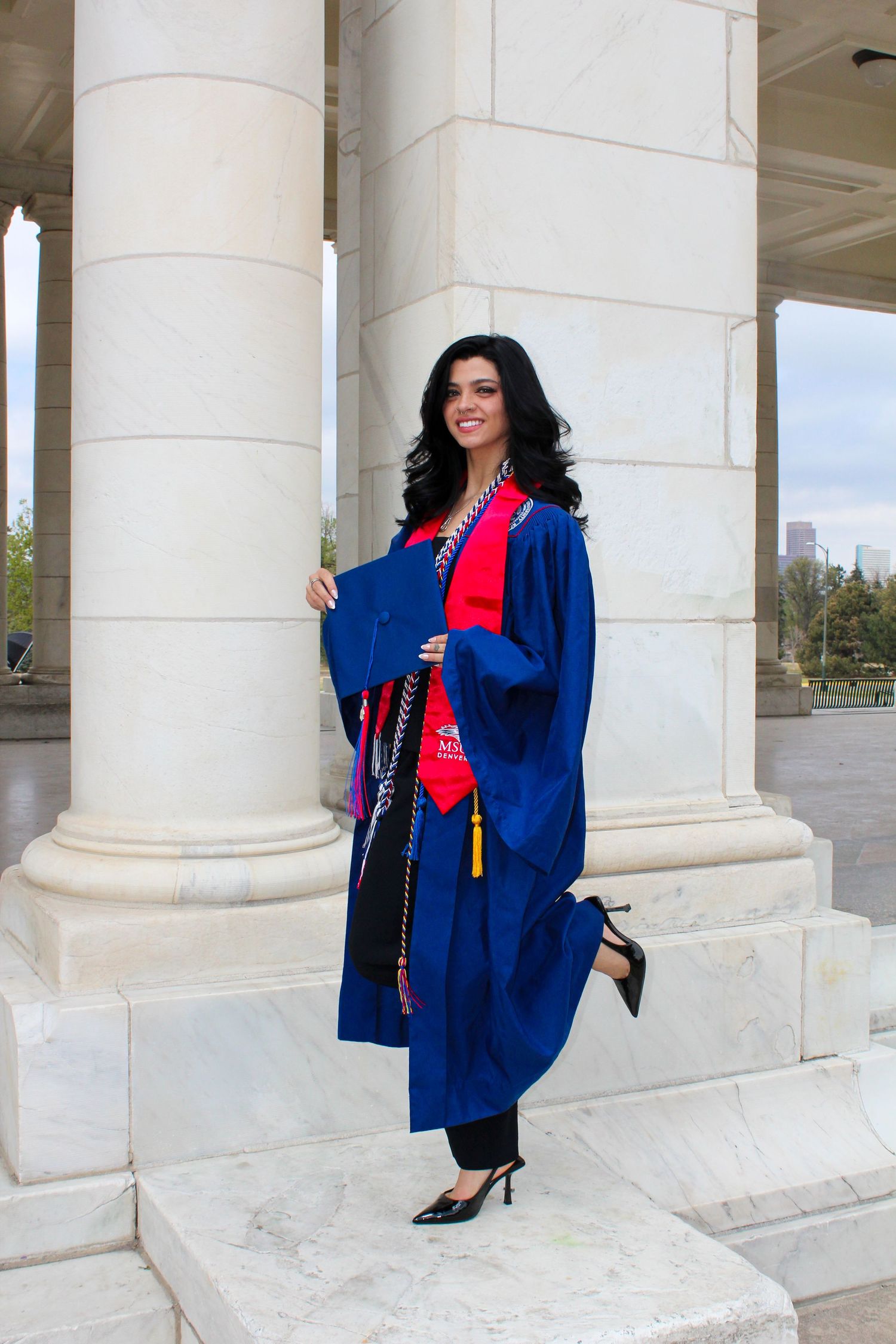 MSU Denver graduate in blue cap and gown with red stole posing between white marble columns outdoors.
