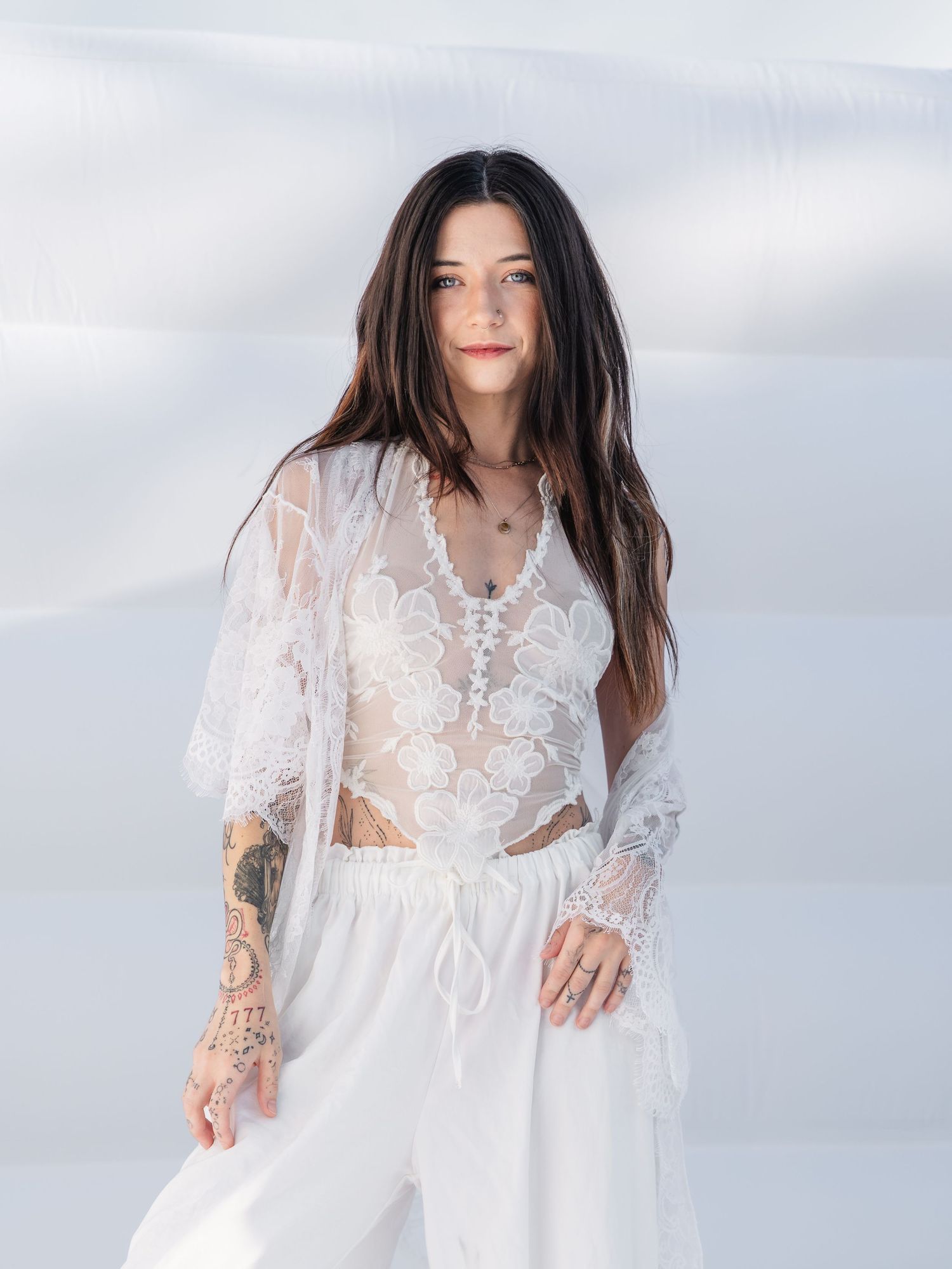 Woman in white lace sheer top and white pants with tattoos, posing against a white background.
