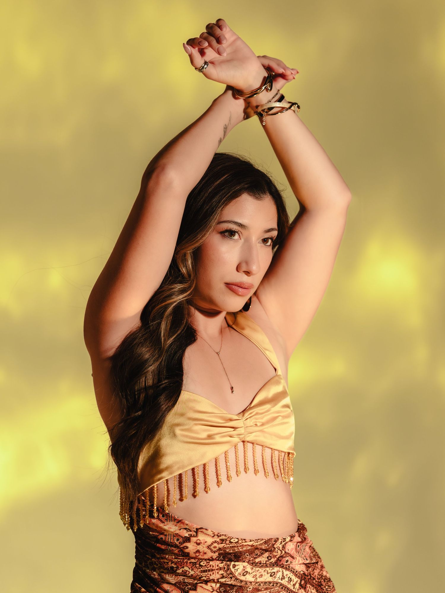 Young woman in gold fringed crop top and sequin skirt poses with arms raised against yellow background.
