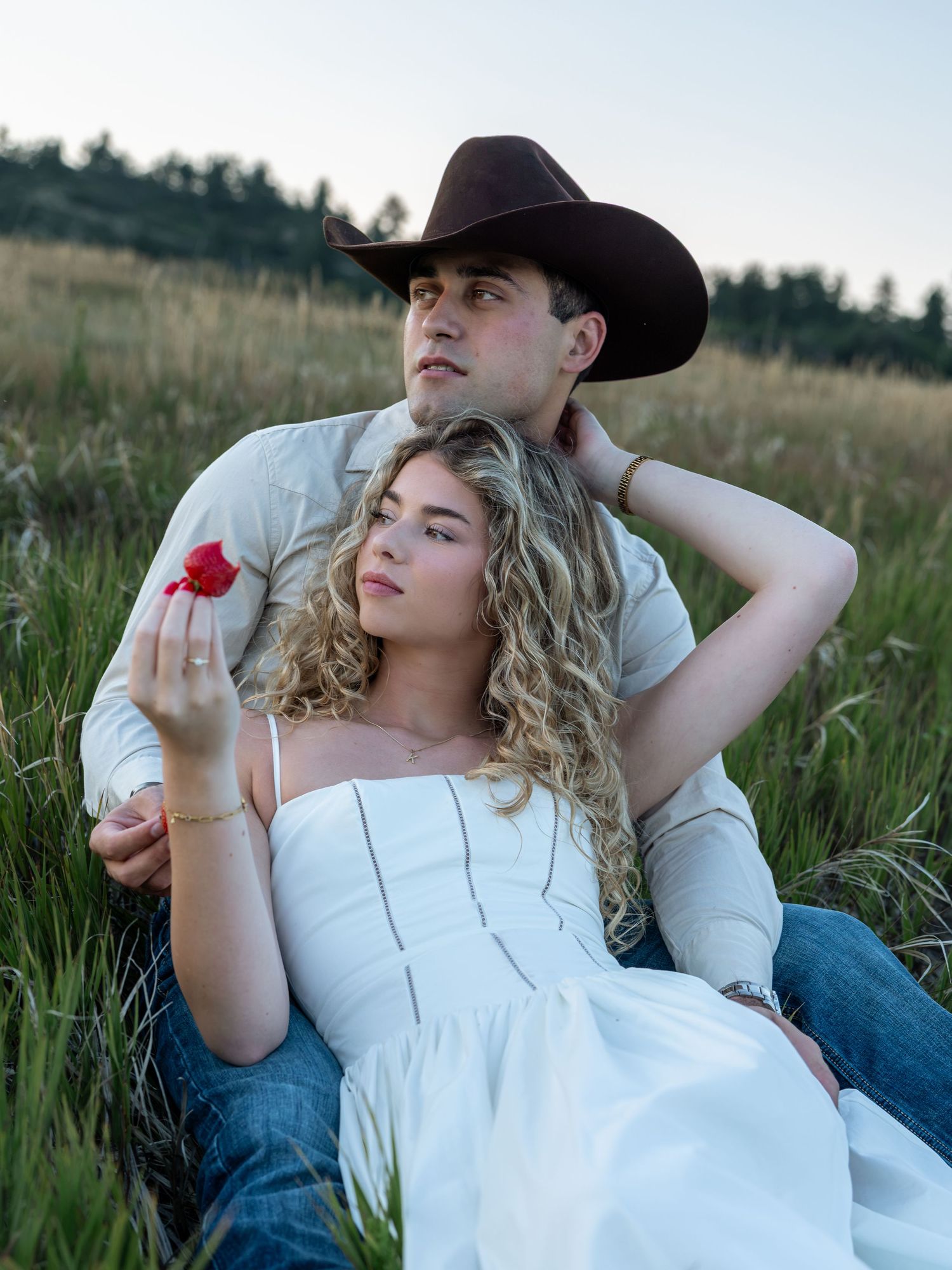 Couple sitting in a green field, woman in white dress holding a red rose, man in cowboy hat embracing her.
