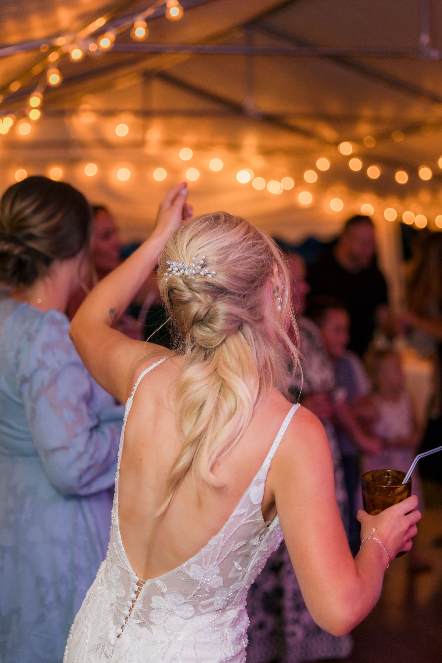 Bride in backless lace gown with floral hair accessory dancing under string lights at wedding reception.