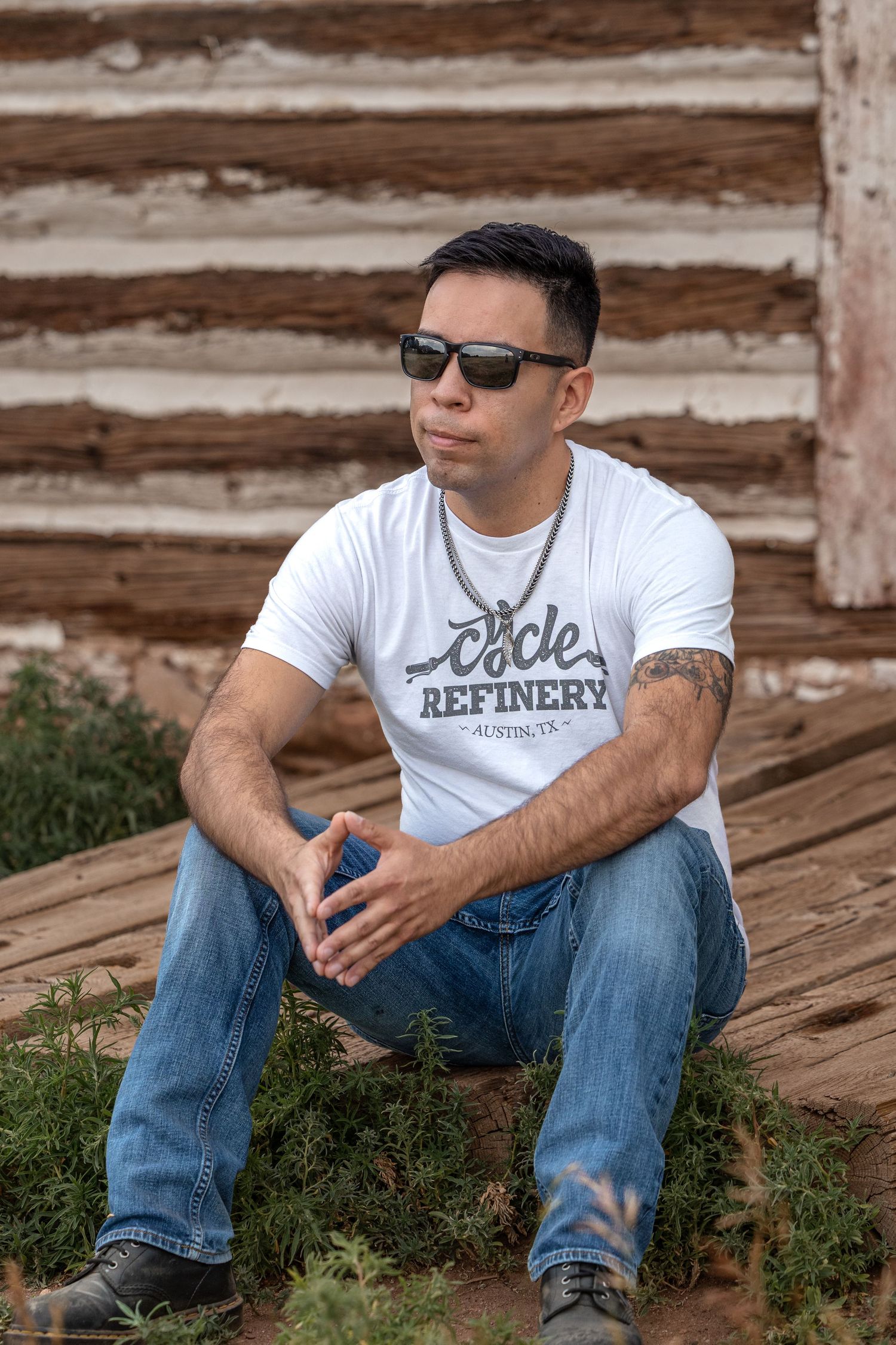 Man in white Cycle Refinery Austin TX t-shirt and sunglasses sitting outdoors near rustic log building.