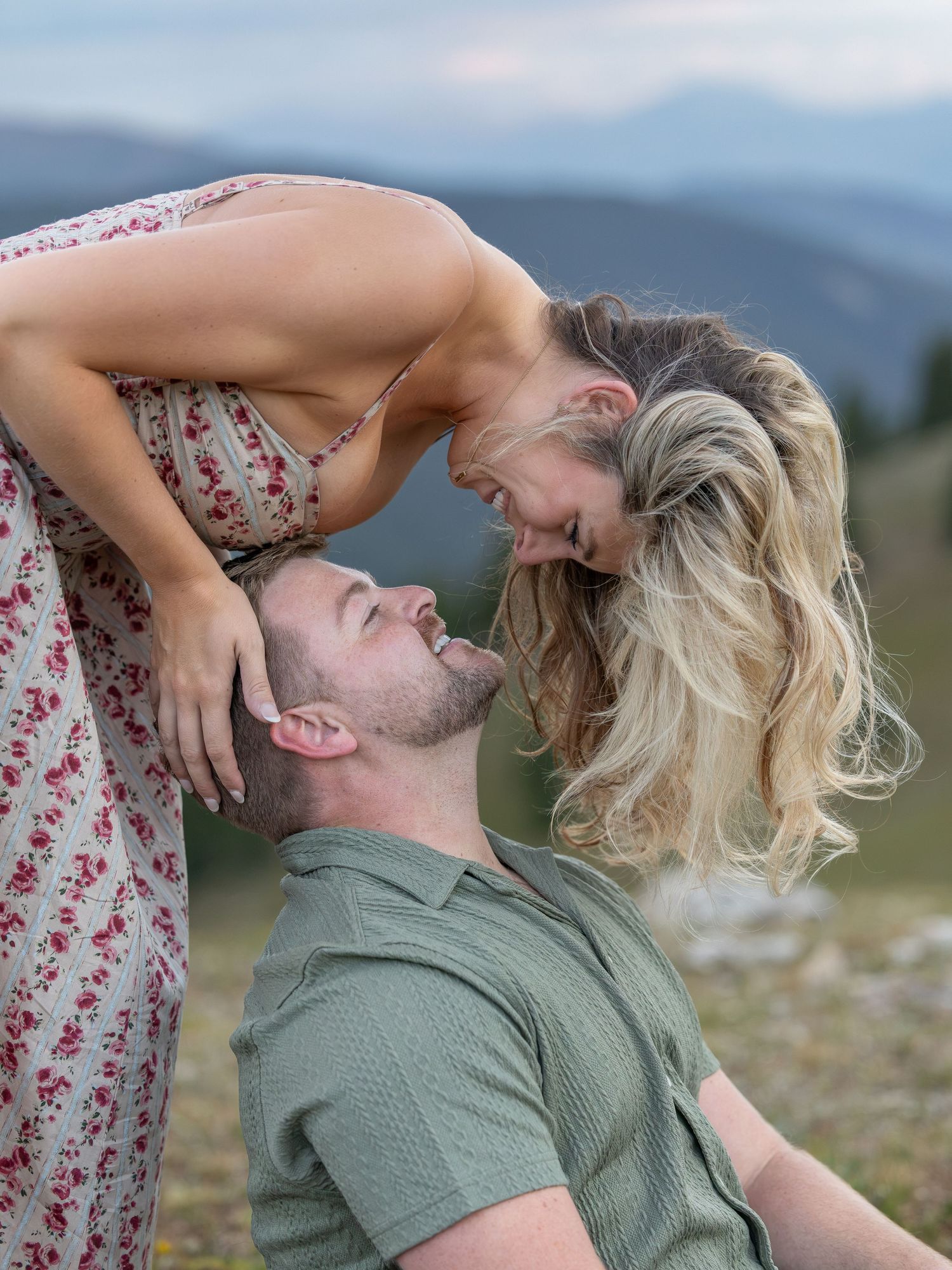 A couple shares a romantic moment outdoors with mountains in the background, woman leaning over man smiling up at her.