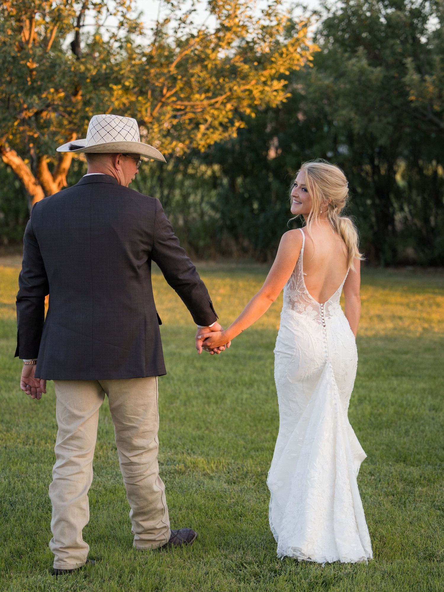 Bride in backless lace gown holds hands with groom in cowboy hat walking through green meadow at golden hour.