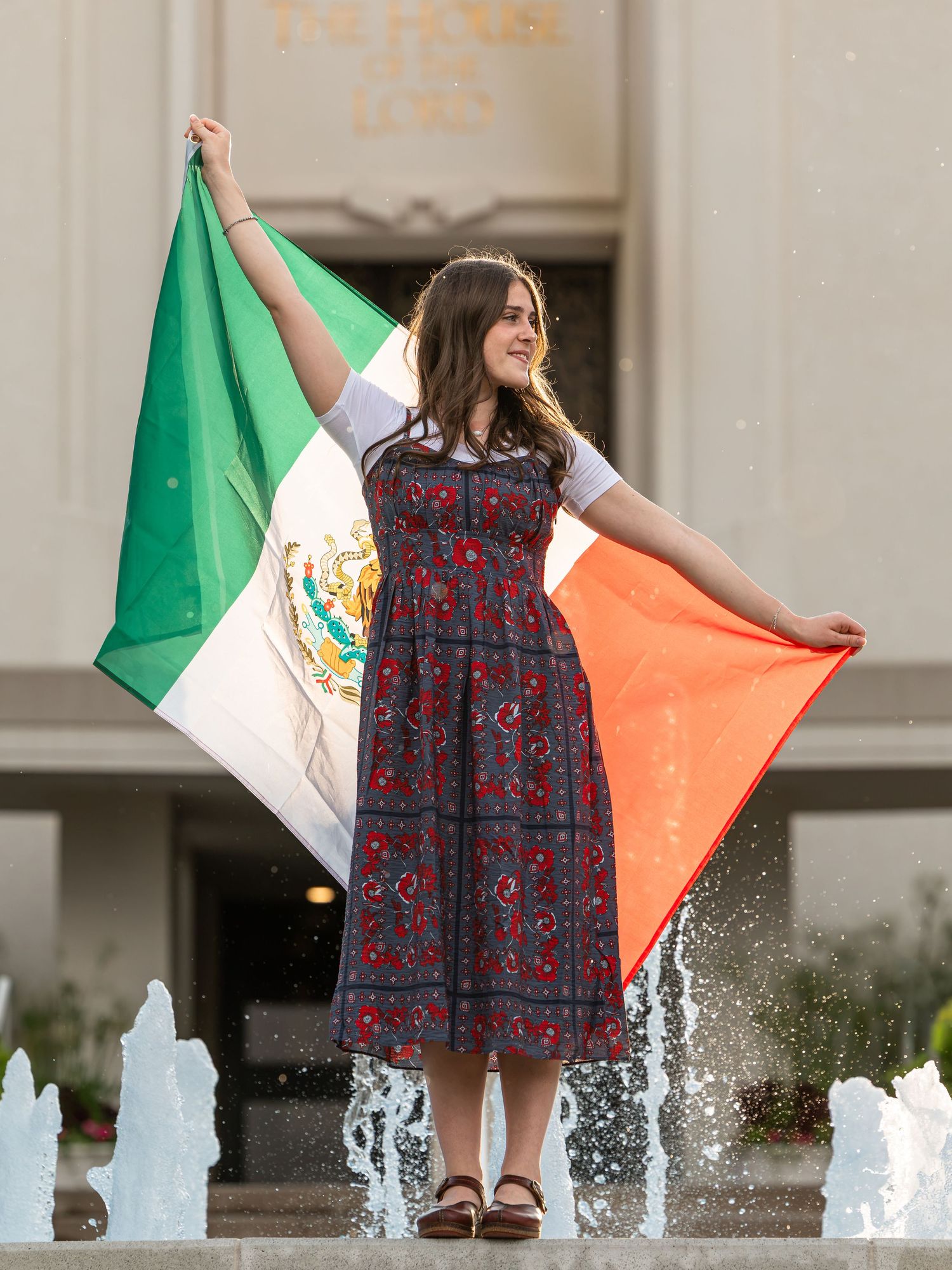 Young woman in floral dress holding Mexican flag by fountain outside building.