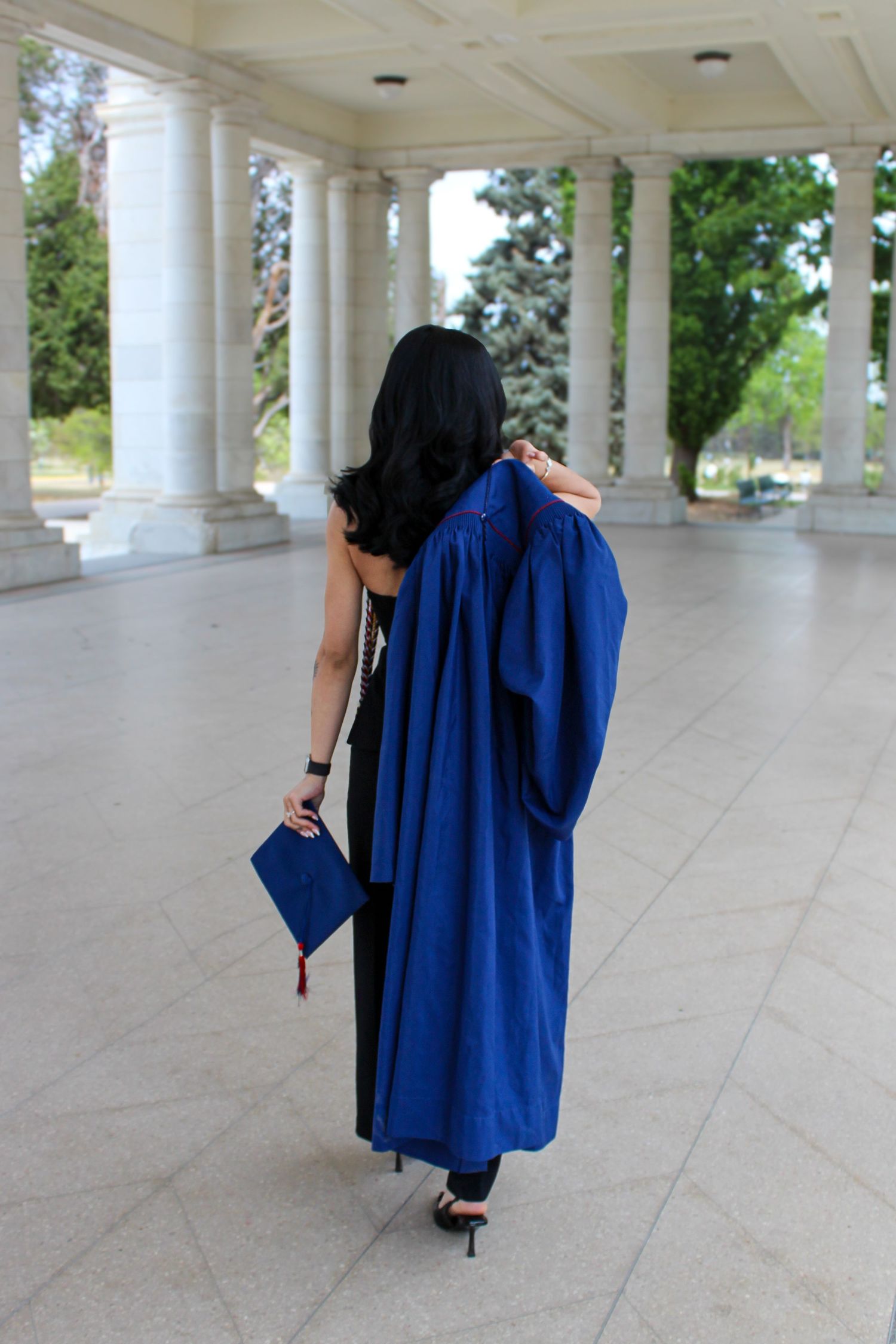 Woman in blue graduation gown and cap walking through elegant columned pavilion, viewed from behind.