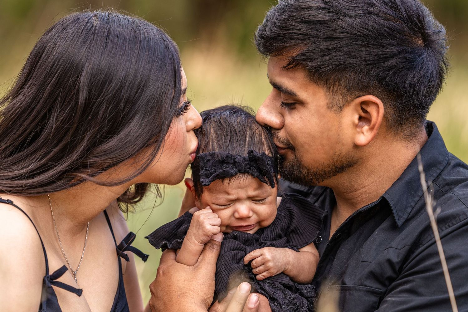 Parents in black outfits kissing their crying newborn baby dressed in black outdoors.