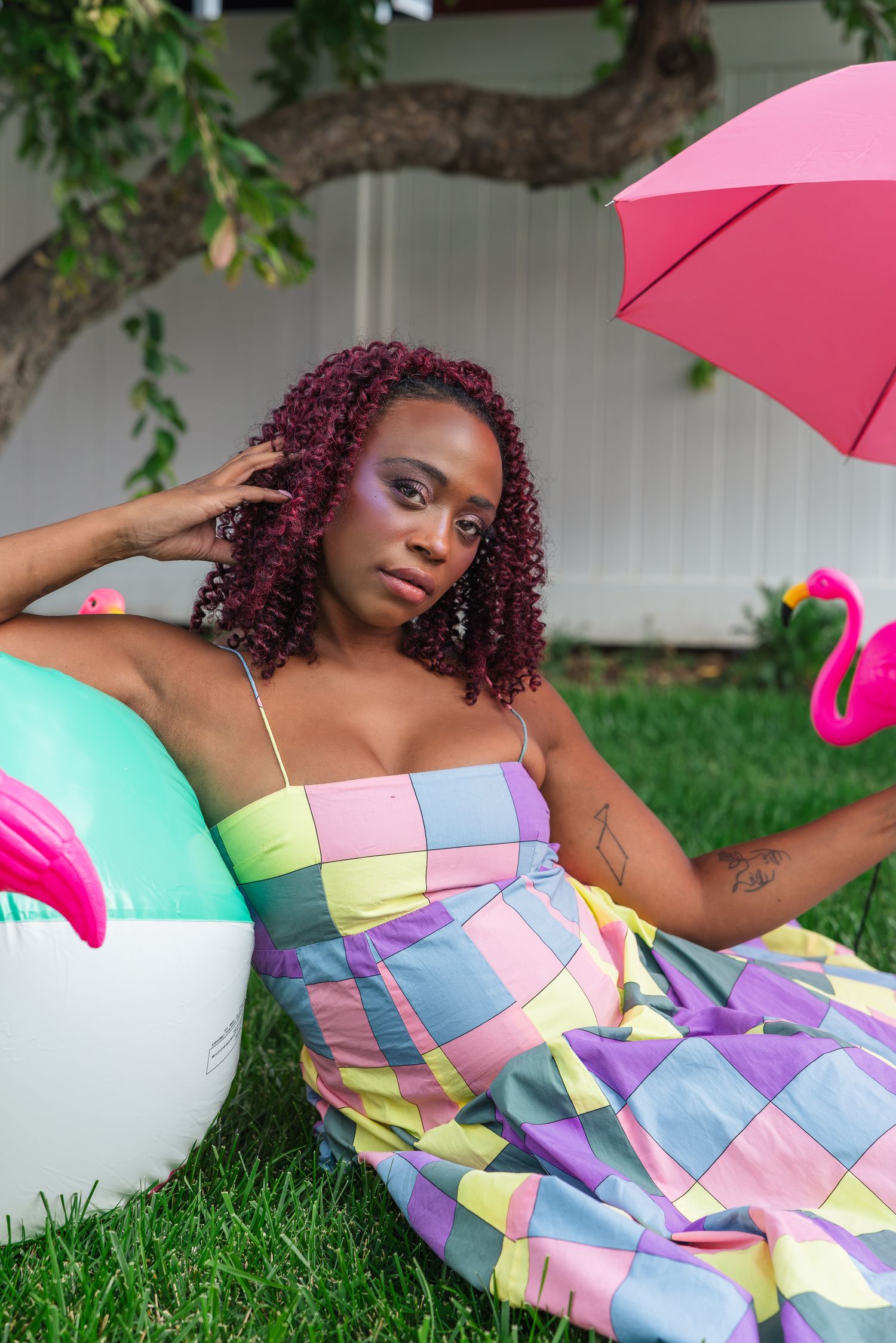 A woman in a colorful plaid dress sits on grass with a flamingo float, beach ball, and pink umbrella.