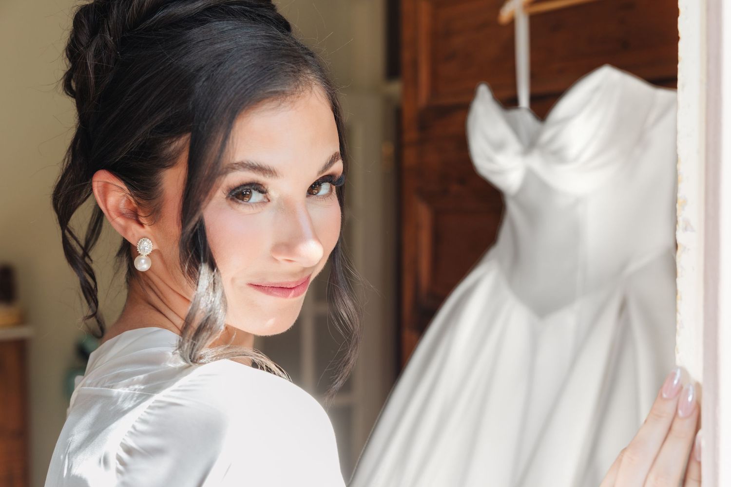 Bride in white satin robe poses beside her elegant strapless wedding gown hanging in a doorway.