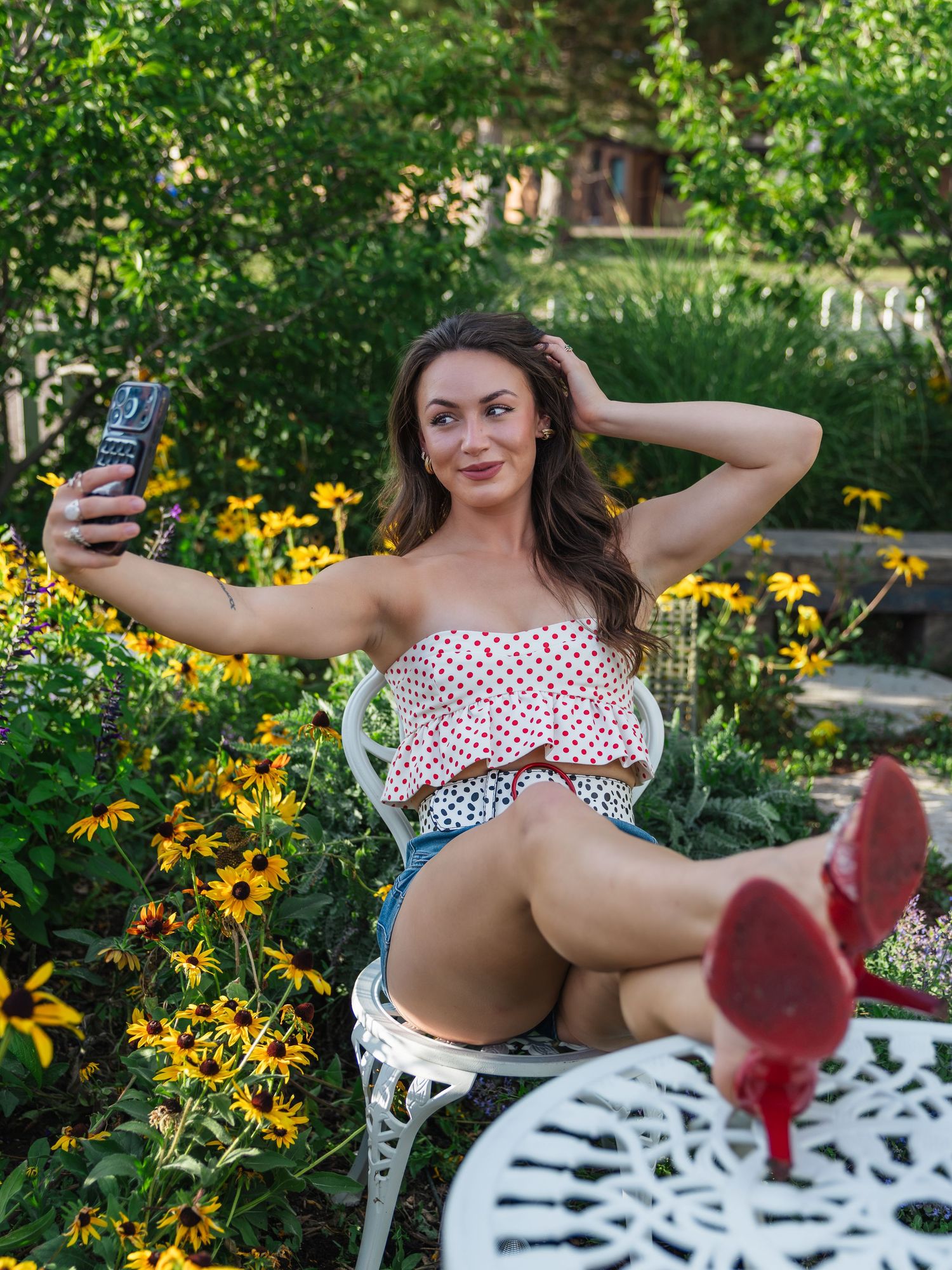 Woman in polka dot top taking selfie outdoors, seated in white garden chair surrounded by yellow flowers.