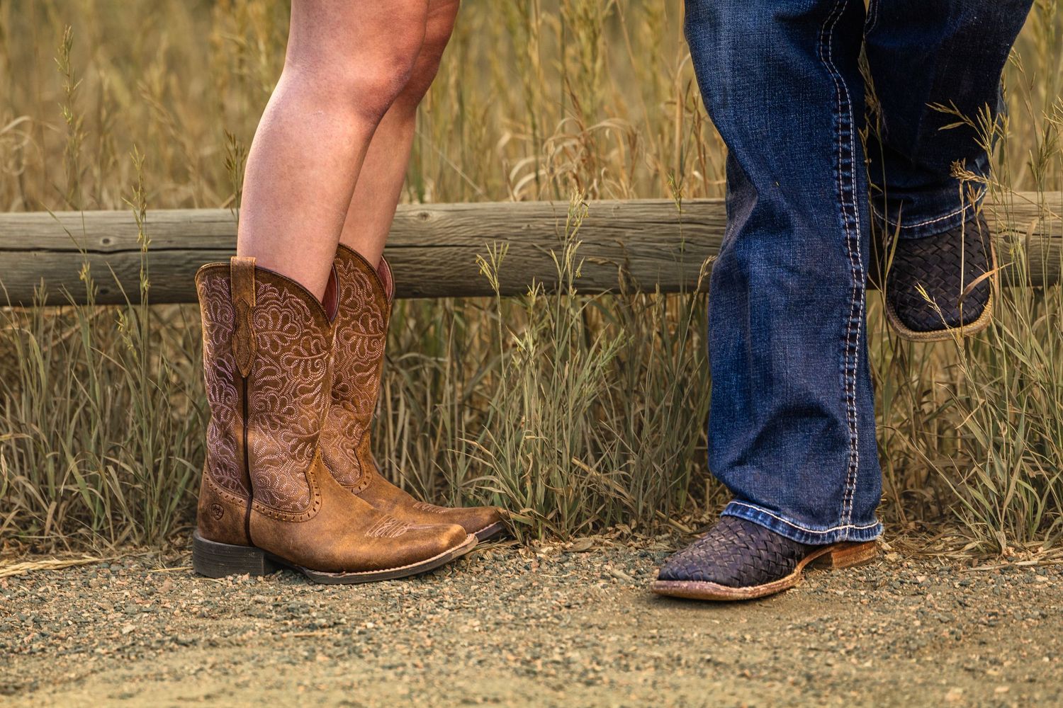 Two people wearing cowboy boots standing on a gravel path beside a wooden fence with tall grass in the background.