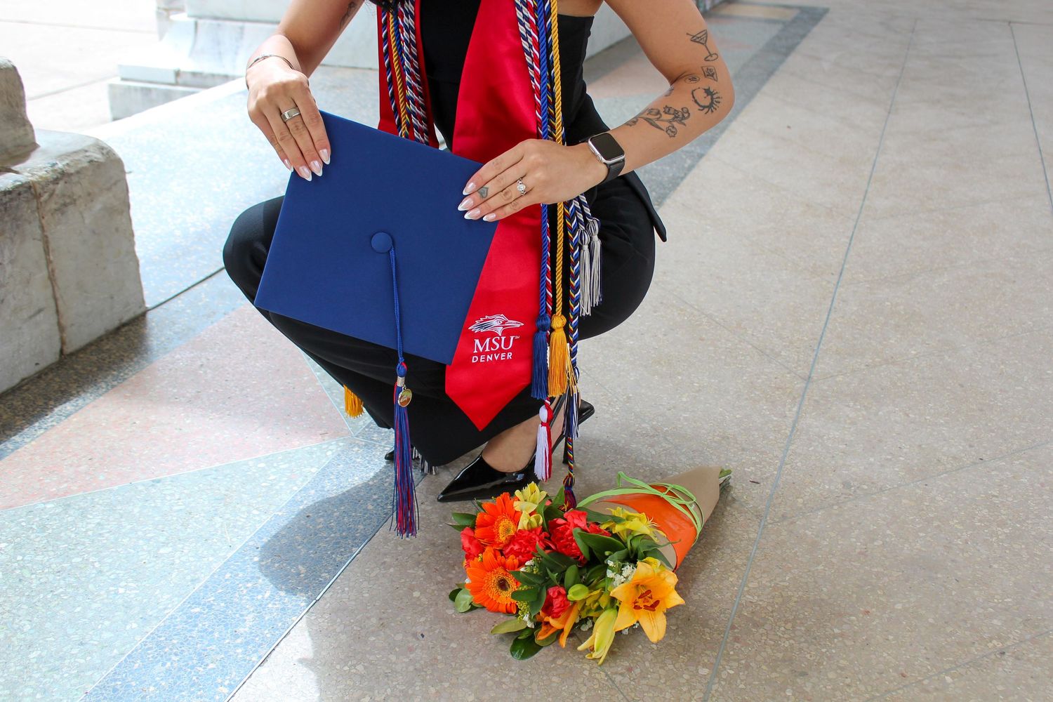 Graduate holds blue MGT cap and red sash while kneeling beside colorful flower bouquet on stone steps.