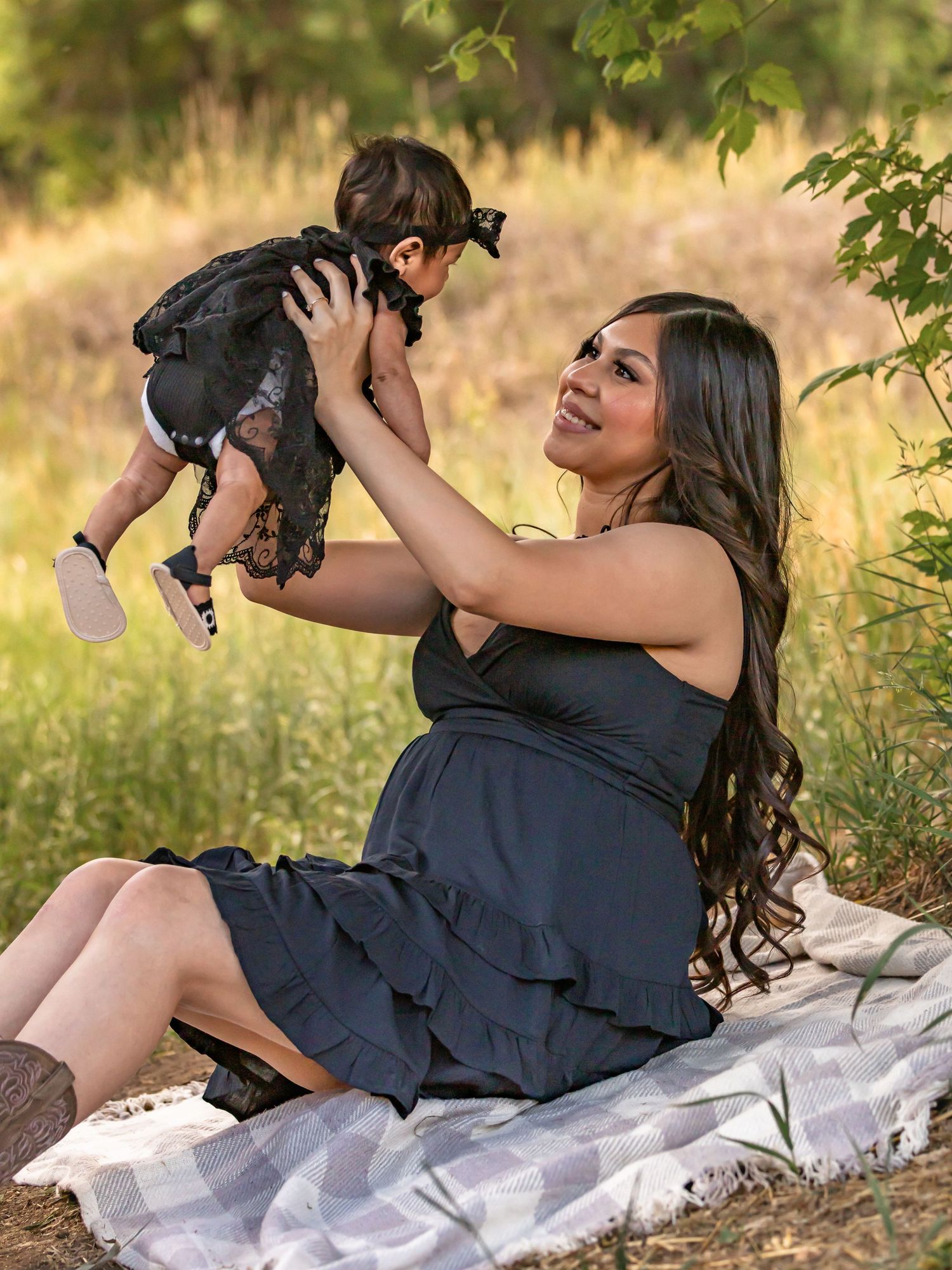 A young woman in a black dress sits outdoors holding up a baby in a black lace outfit, smiling joyfully.