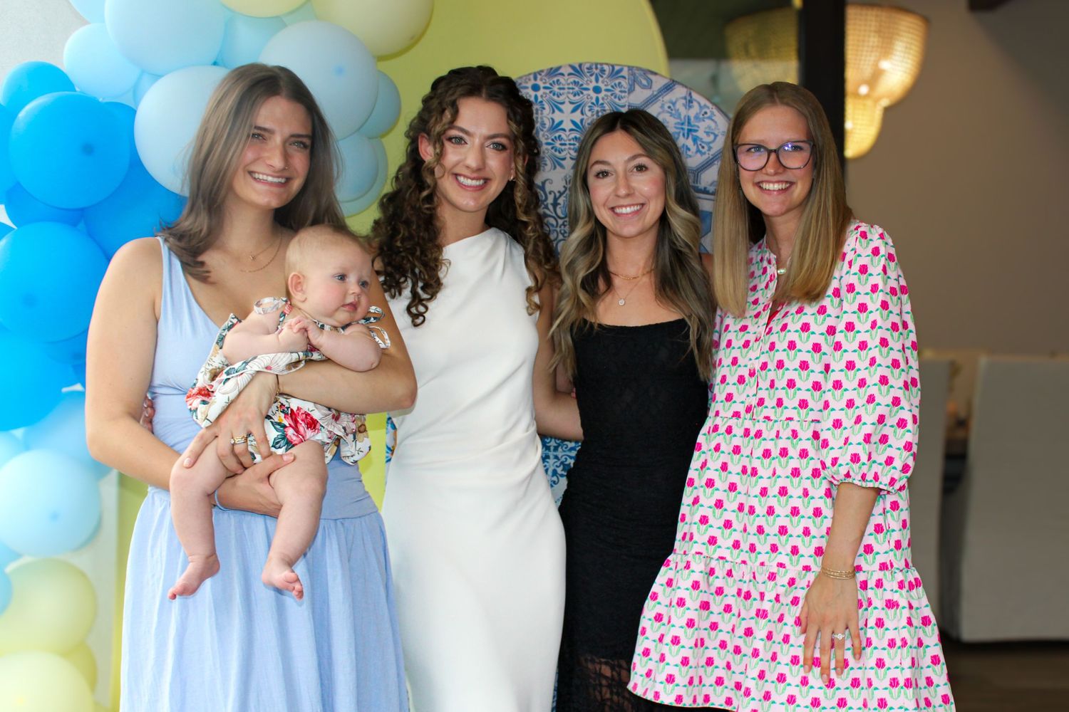 Four women smiling at a baby shower, one holding an infant, surrounded by blue and yellow balloon decorations.