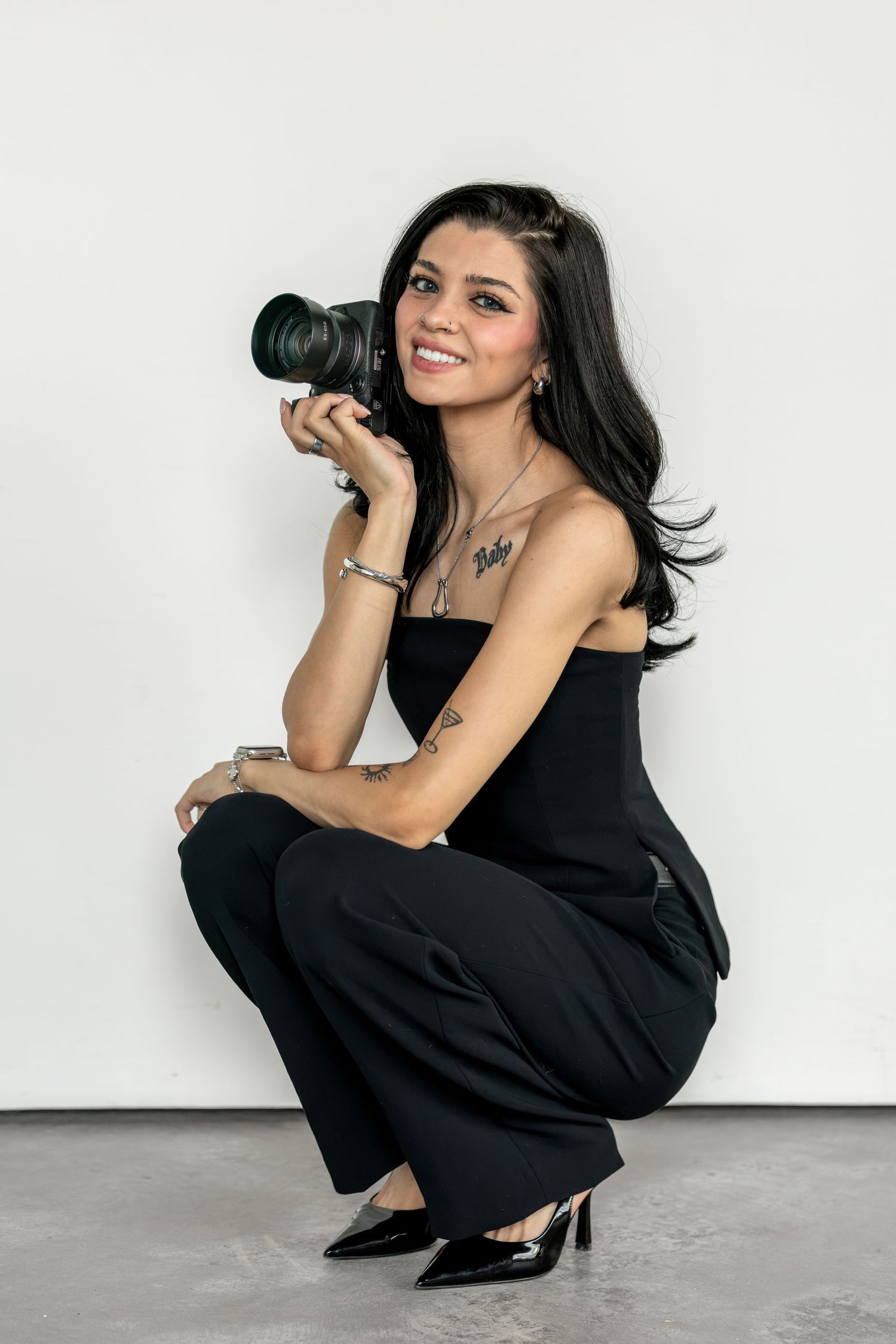 Young woman in black jumpsuit crouching while holding a DSLR camera, smiling against a white wall.